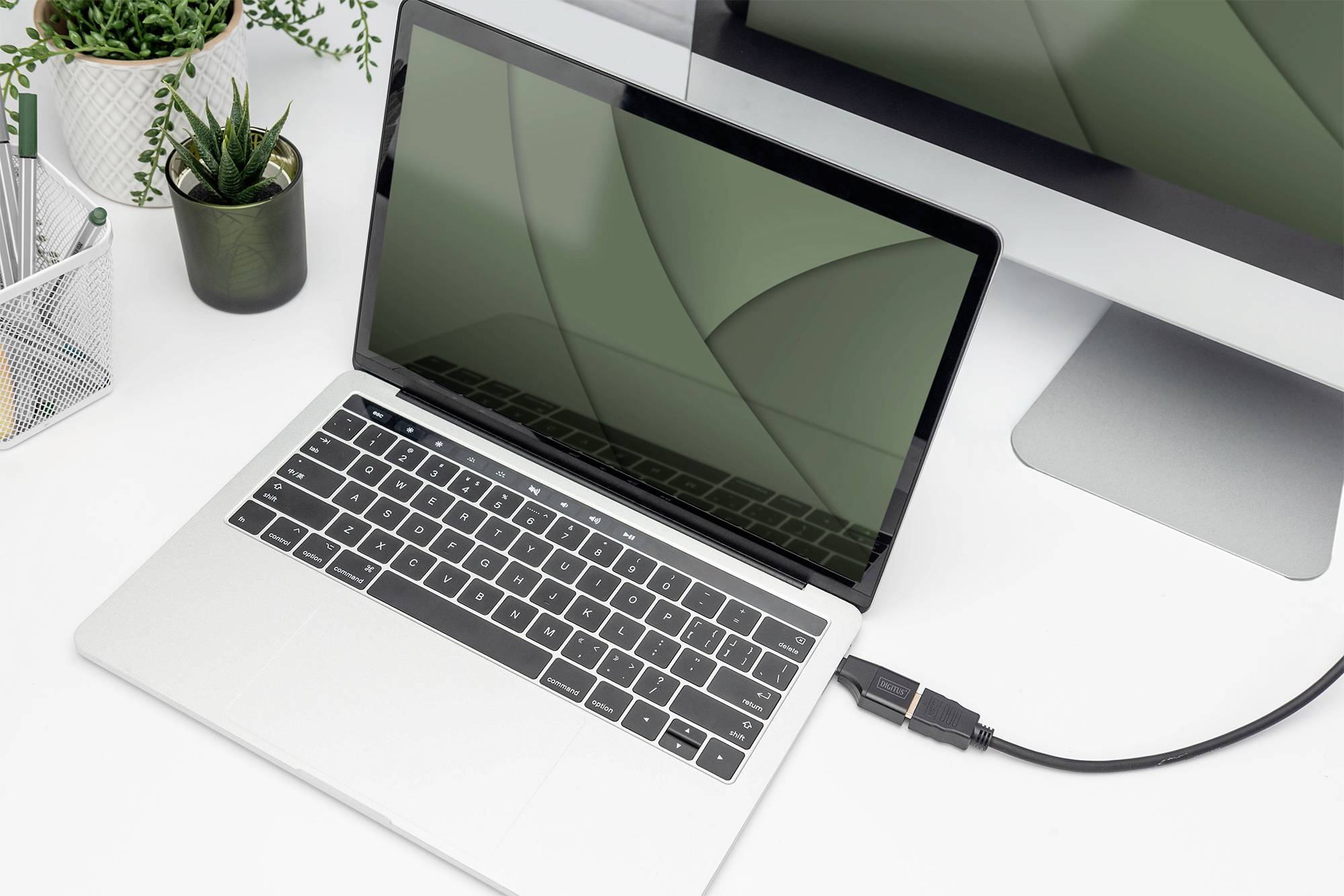 En bærbar computer på et skrivebord er tilsluttet en større skærm via et HDMI-kabel. Til højre står en lille plante.