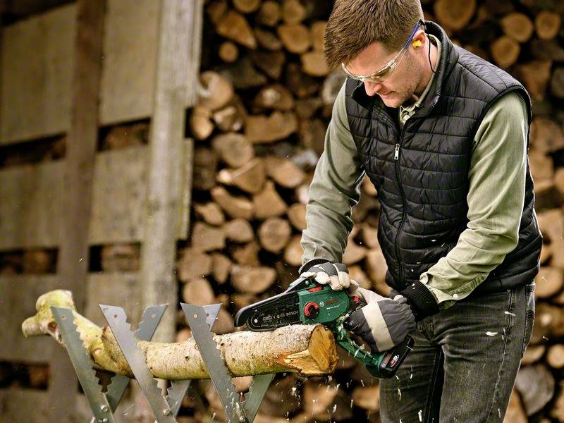 En mand save et træstykke med en elektrisk kædesav i et værksted. En stak brænde ses i baggrunden.