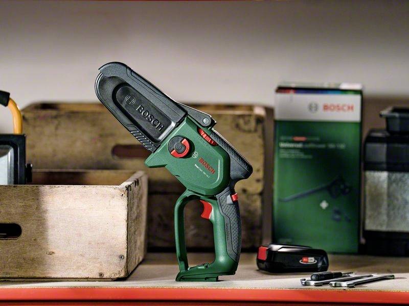 En kompakt grøn elektrisk kædesav fra Bosch står i et værksted, omgivet af træ-kasser og værktøjstilbehør.
