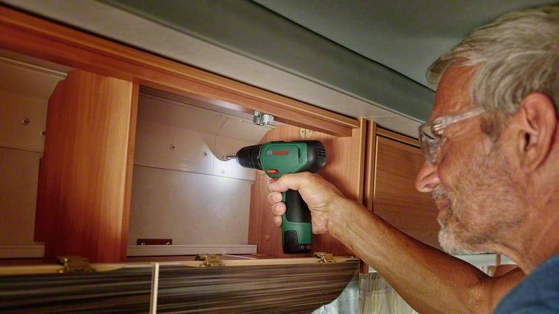 En mand borer med et elektrisk værktøj i et træskab i campingvognen, iført sikkerhedsbriller og fokuseret på sit arbejde.