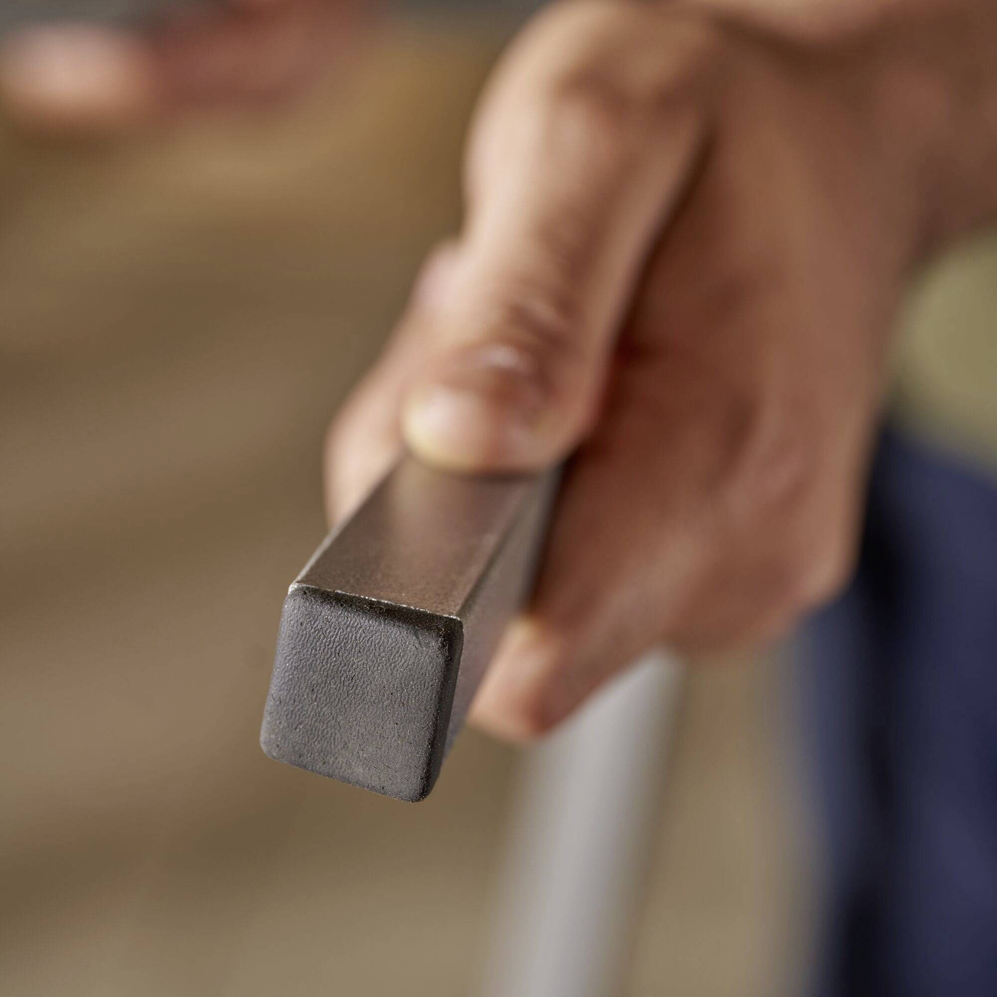 En hånd holder fast om et metalstykke med et firkantet tværsnit. Baggrunden er sløret.