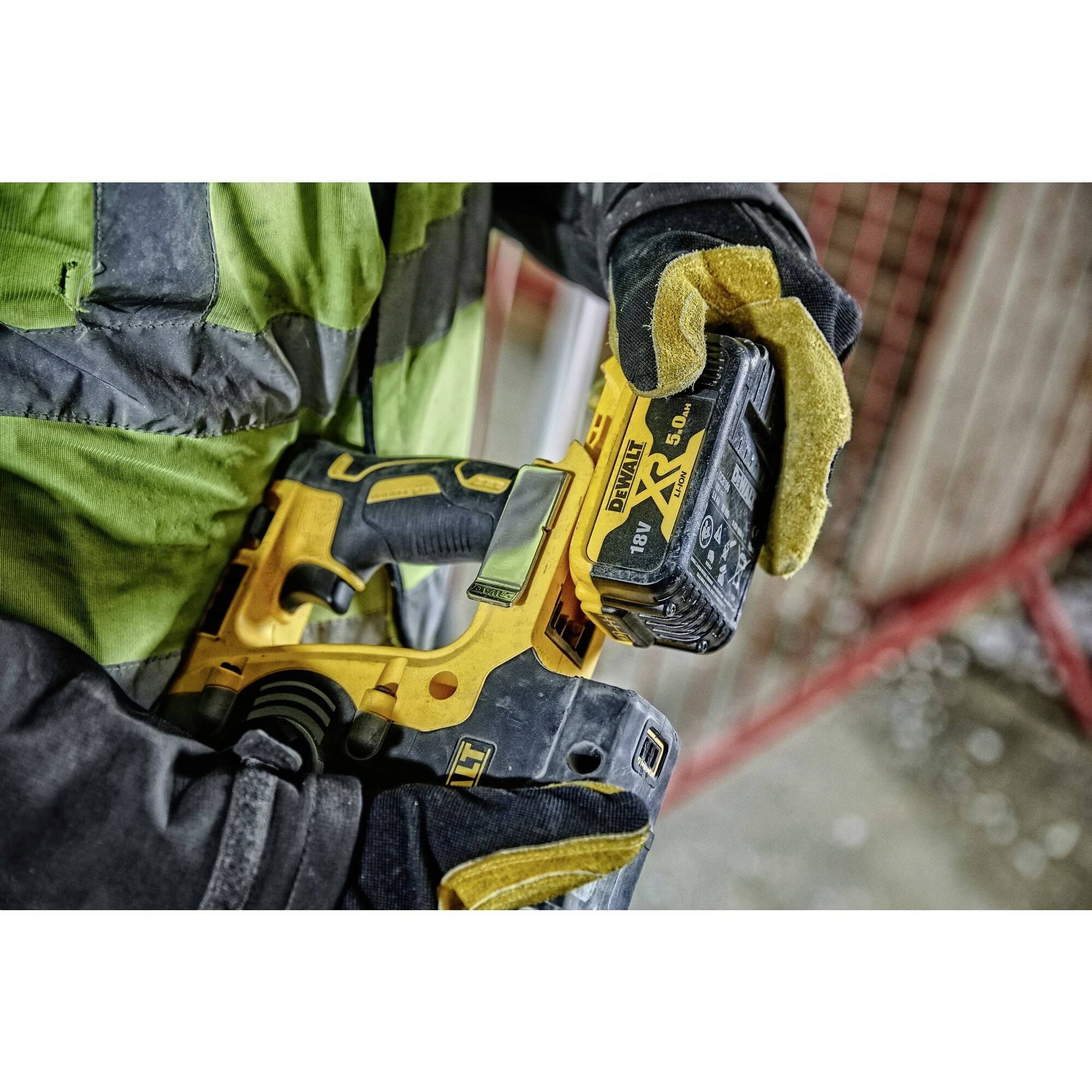 En person bærer handsker og holder en gul elektrisk håndboremaskine, klar til brug på en byggeplads.