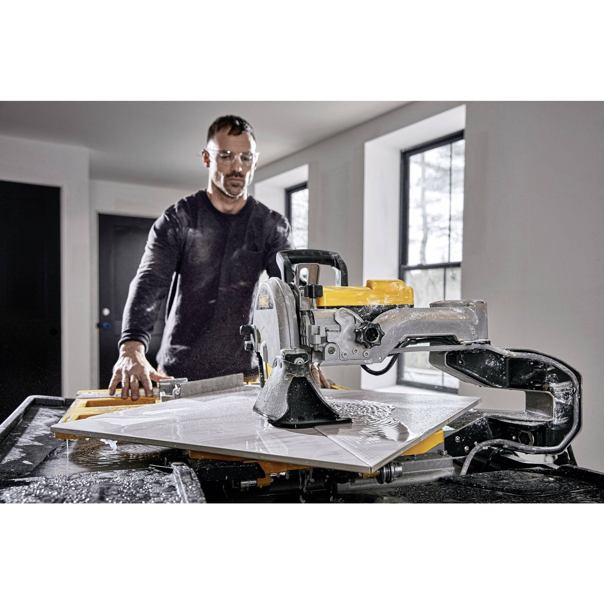 En mand skærer en stor keramisk flise ved hjælp af en gul fliseværktøj. I baggrunden ses vinduer og døre.