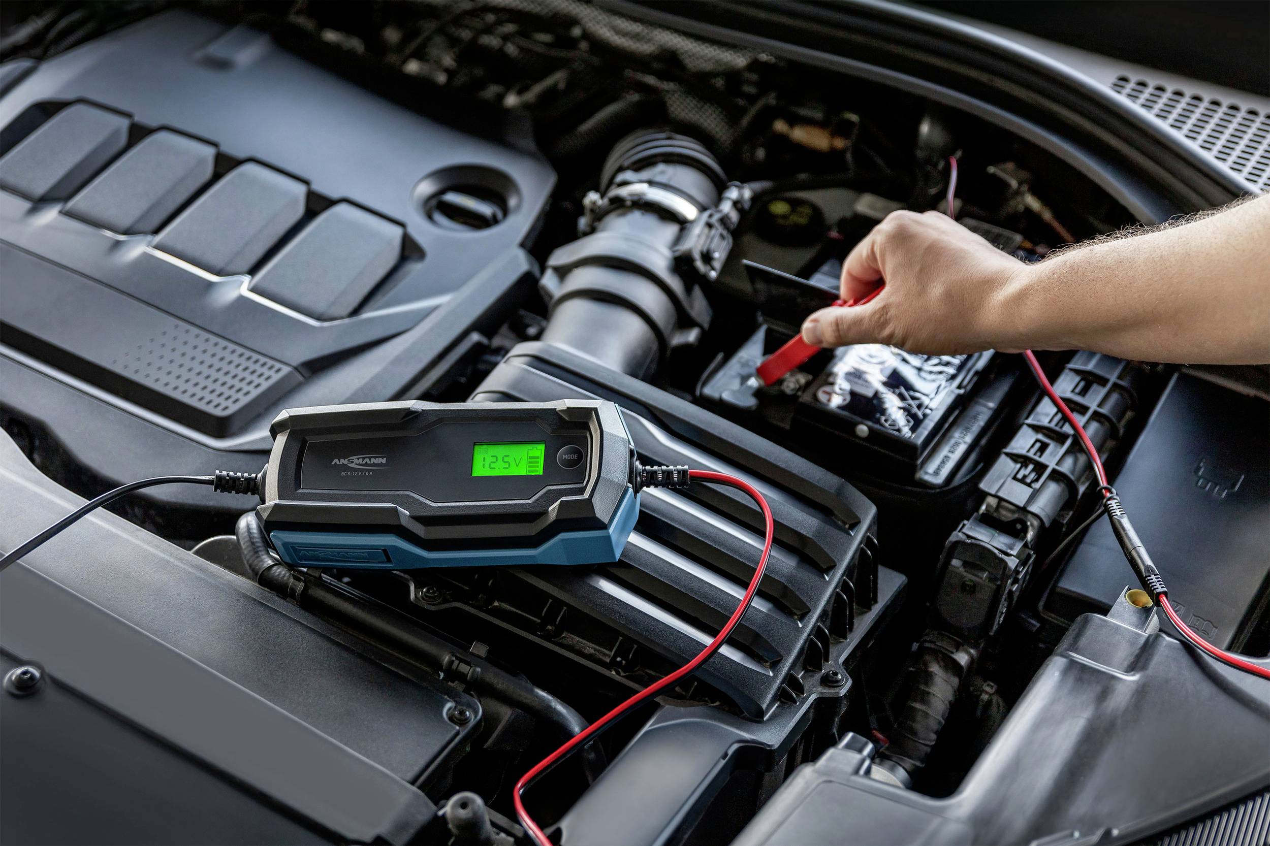 En hånd tilslutter et kabel fra et batteriopladeren til en bilbatteri. Displayet på opladeren viser 12,5V.