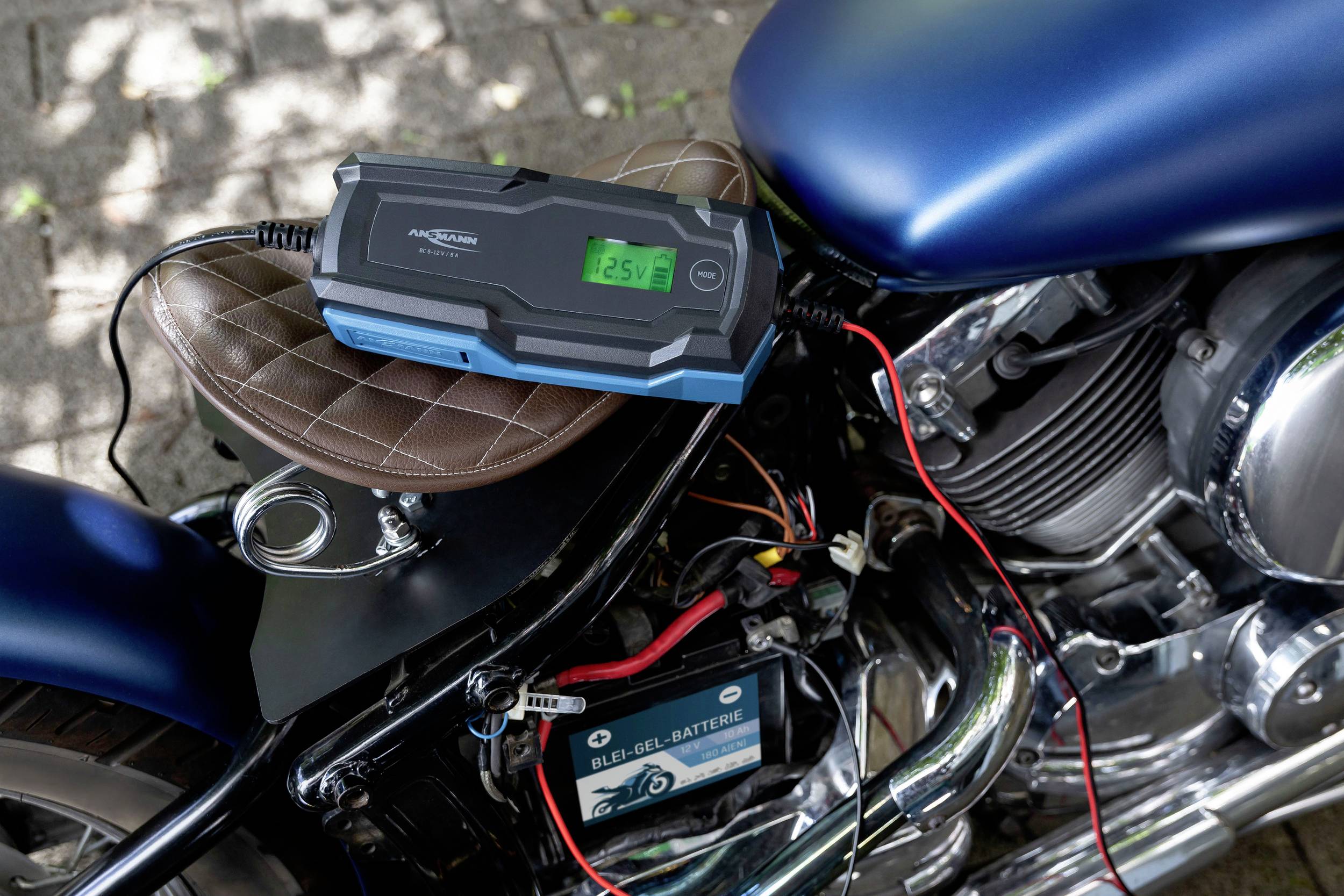 En motorcykel står på et fortov, forbundet med en batterilader. Displayet viser '12.5V'. Skygger fra træer i baggrunden.