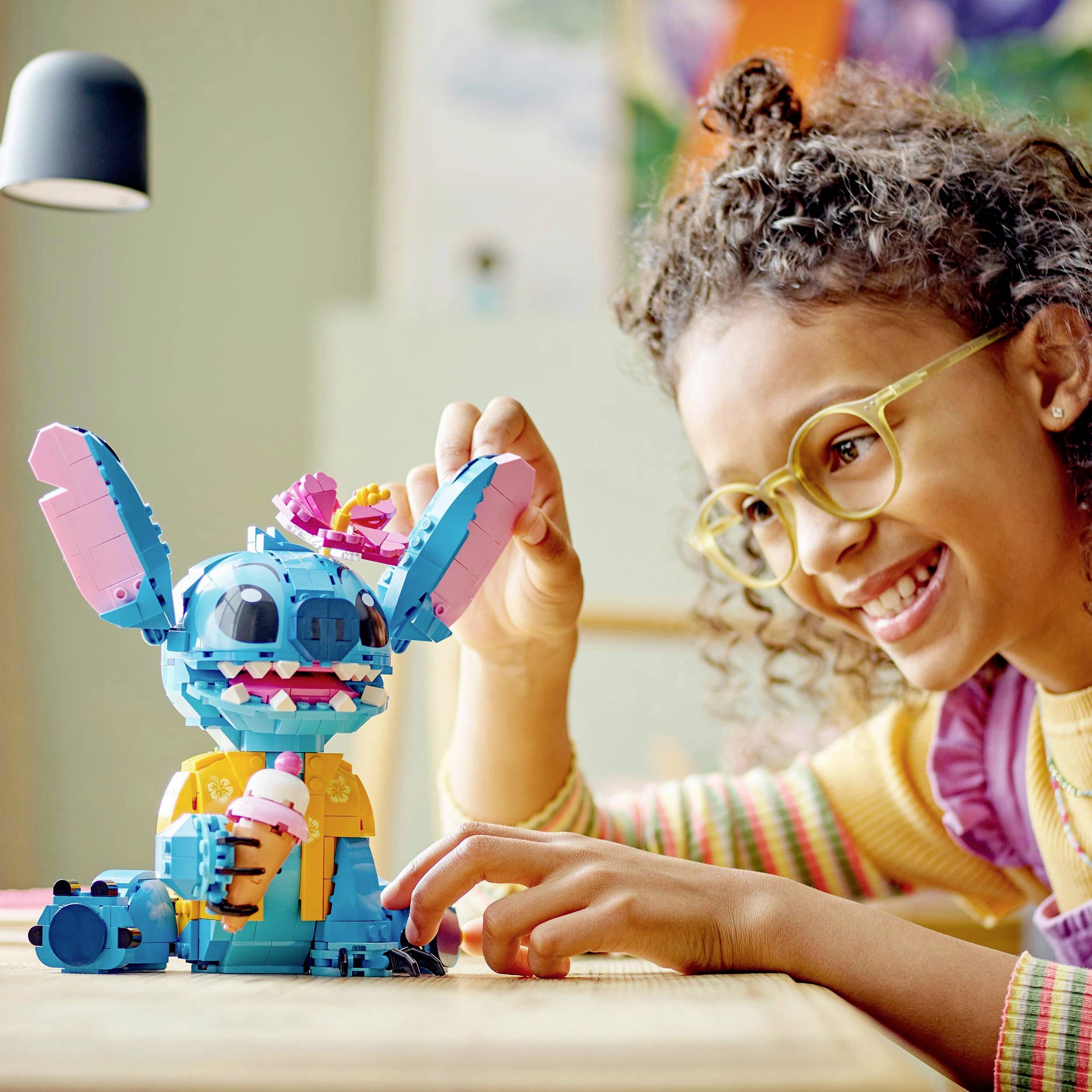 Et pigebarn med briller smiler og peger på en farverig legetøjsfigur på et bord. Figuren holder en lyserød isskål.