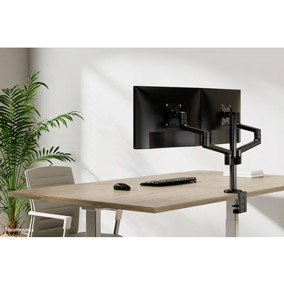 To monitorer på justerbare holders på et skrivebord med tastatur og mus, ved siden af en kontorstol og en plante.
