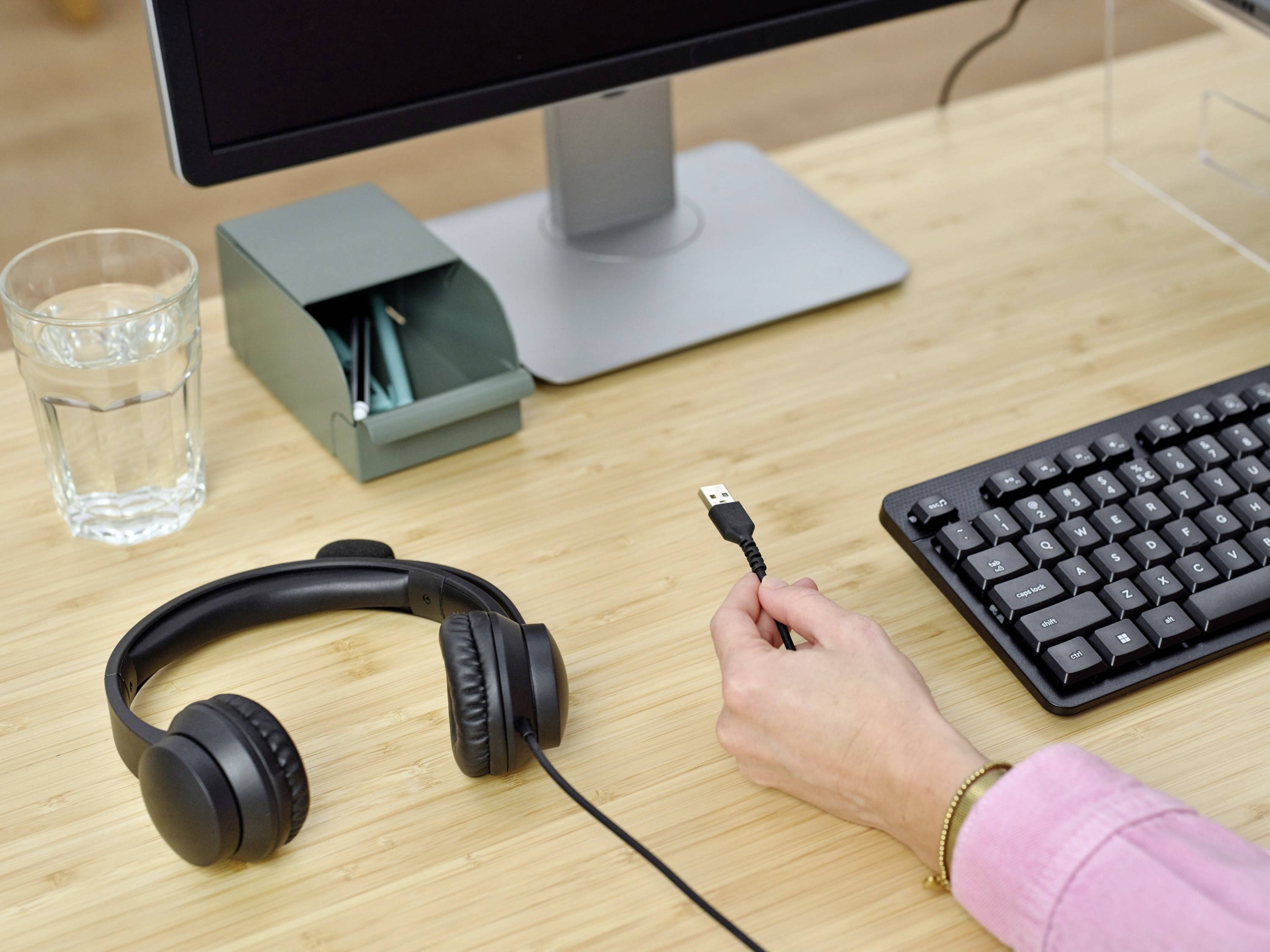 En person holder en USB-stik foran en computerskærm og et tastatur. På bordet ligger også et glas vand og et par høretelefoner.