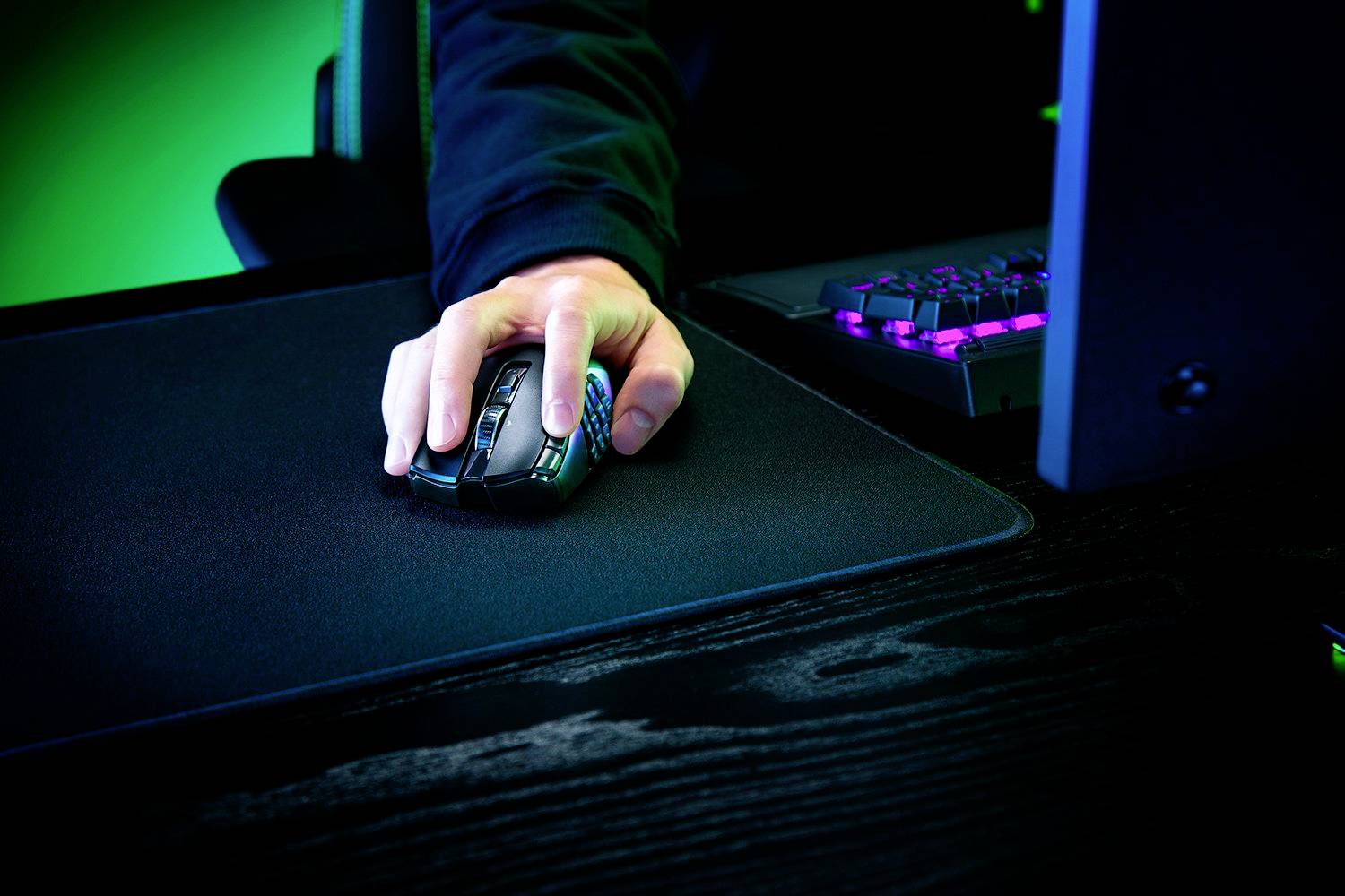 En person bruger en gaming-mus på et musemåtte ved siden af et belyst tastatur.
