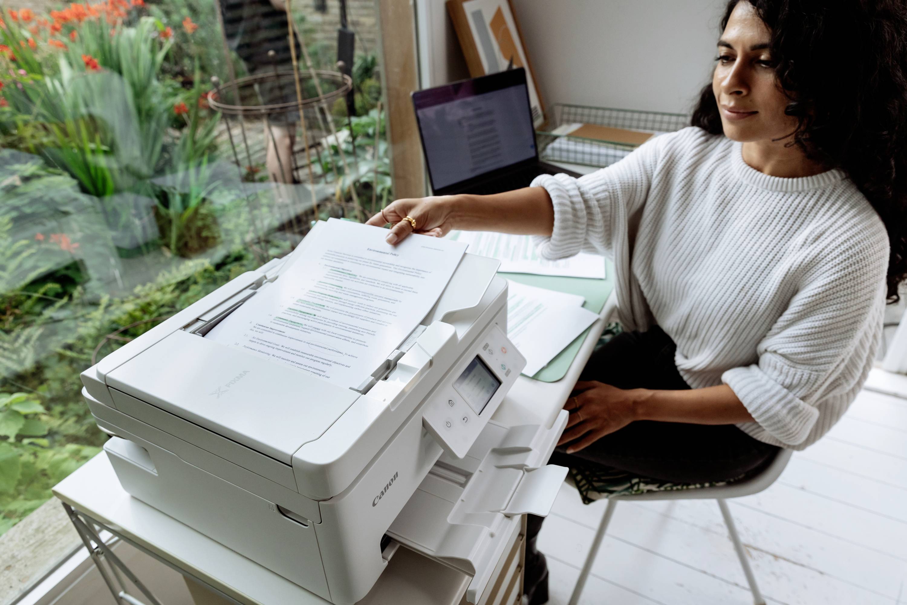 En kvinde bruger en printer i et kontor med udsigt til en have. Hun holder et ark papir. En bærbar computer står på bordet.