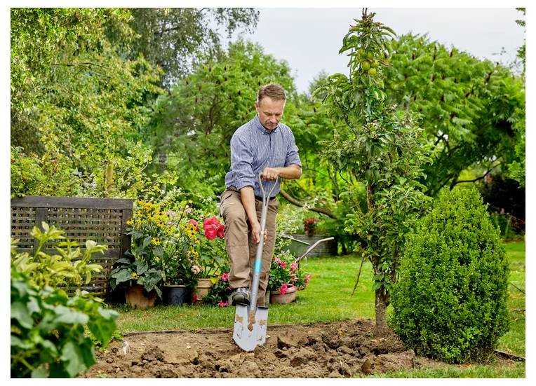 En person i en have er ved at plante et træ med en skovl. Scenen er frodigt grøn med forskellige planter og blomster, hvilket antyder et fokus på havearbejde.