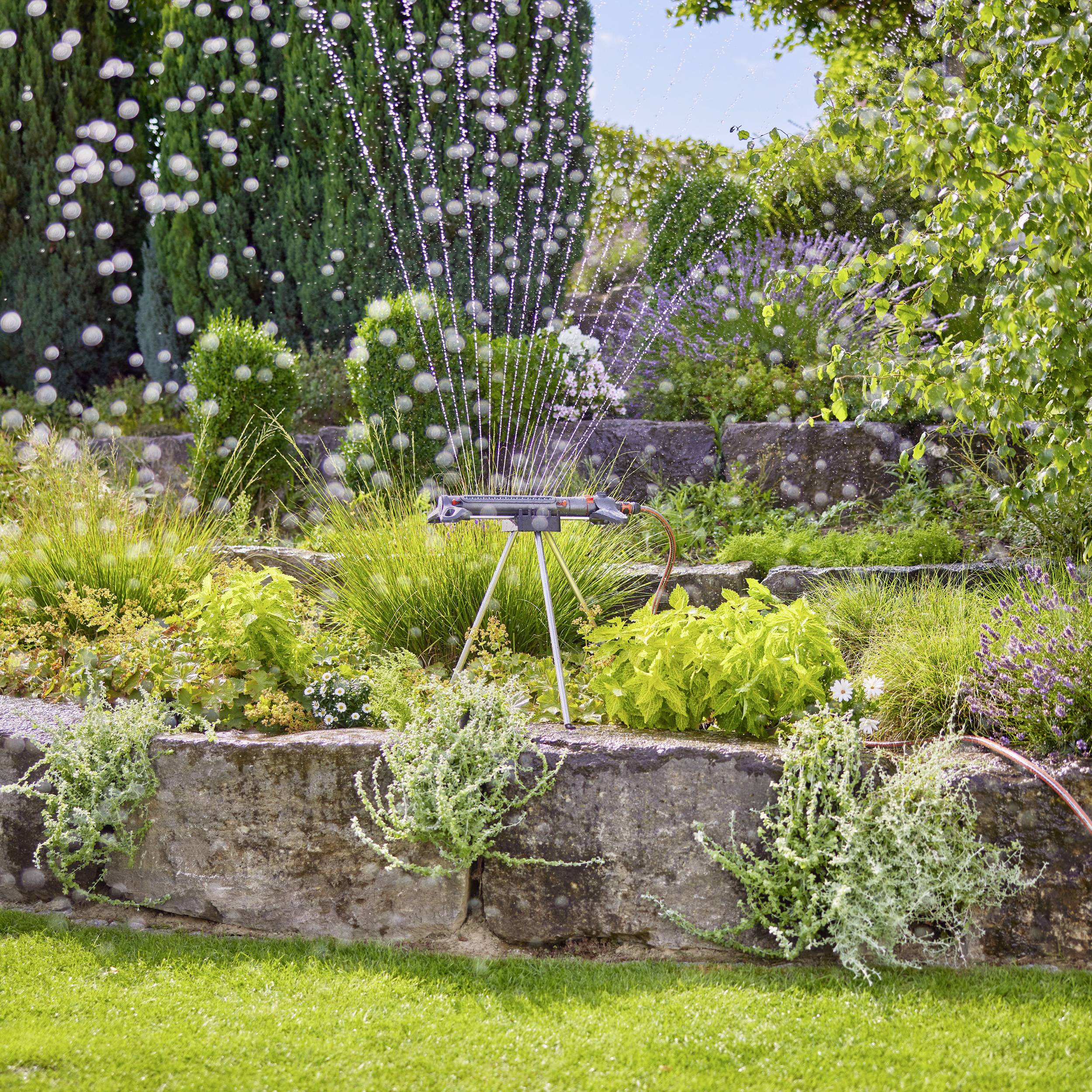 En moderne sprinkler vander en grøn have med blomster og buske. Vanddråber i luften skaber en forfriskende atmosfære.