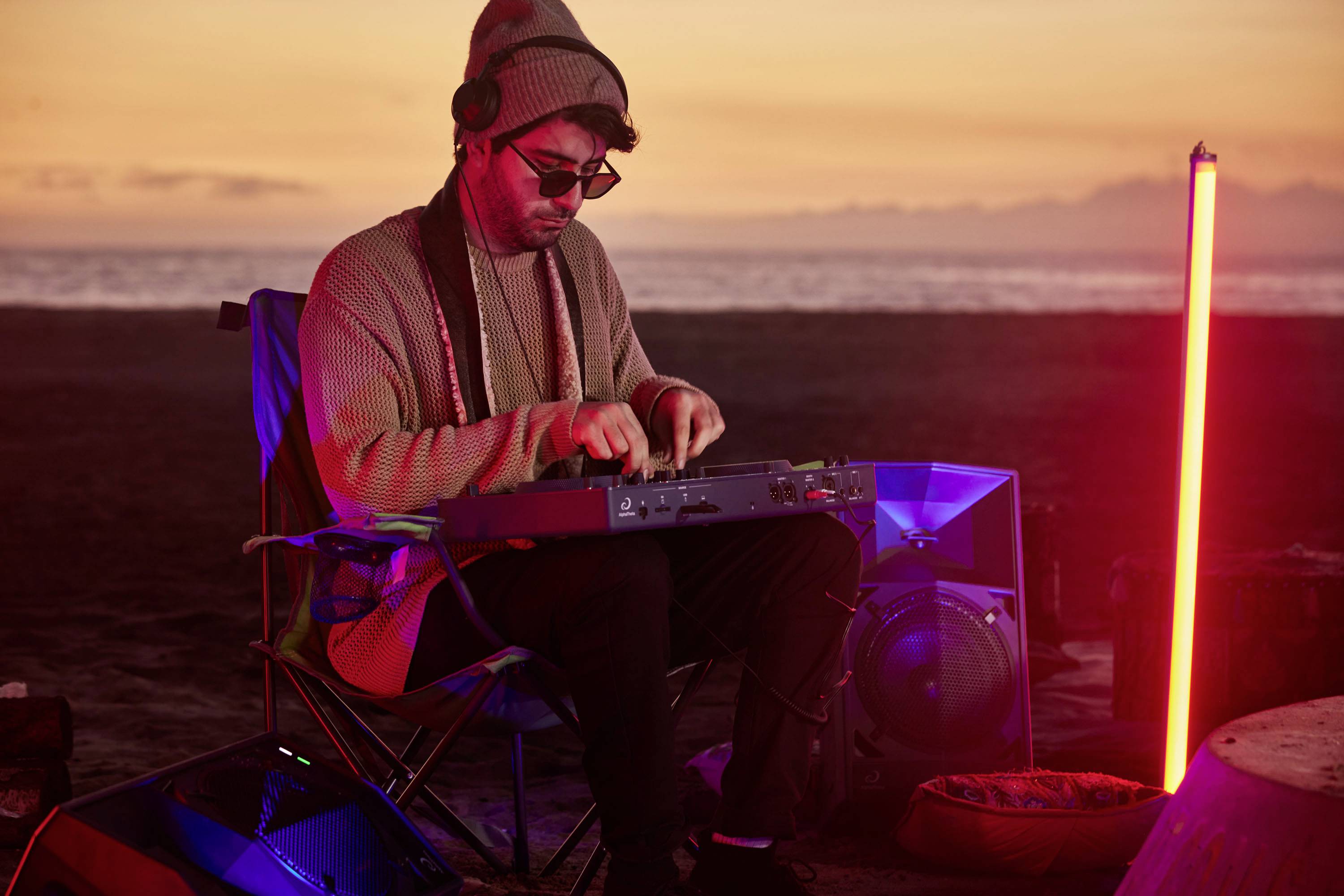 En mand med hovedtelefoner og solbriller sidder og betjener et musikmixerpult ved stranden ved solnedgang.