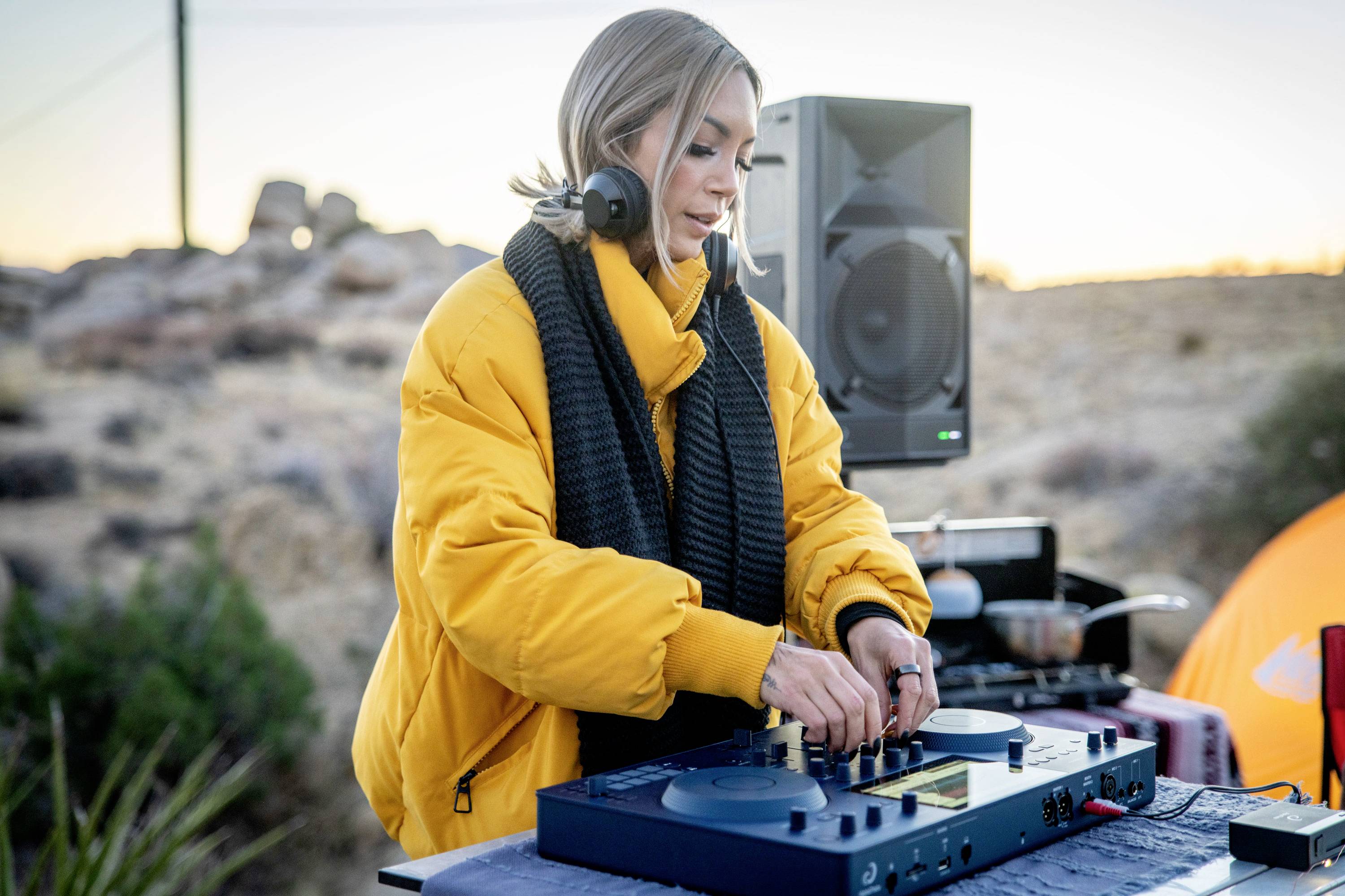 En person i en gul jakke blander musik ved et DJ-bord udendørs ved solnedgang. I baggrunden ses højttalere og telte.