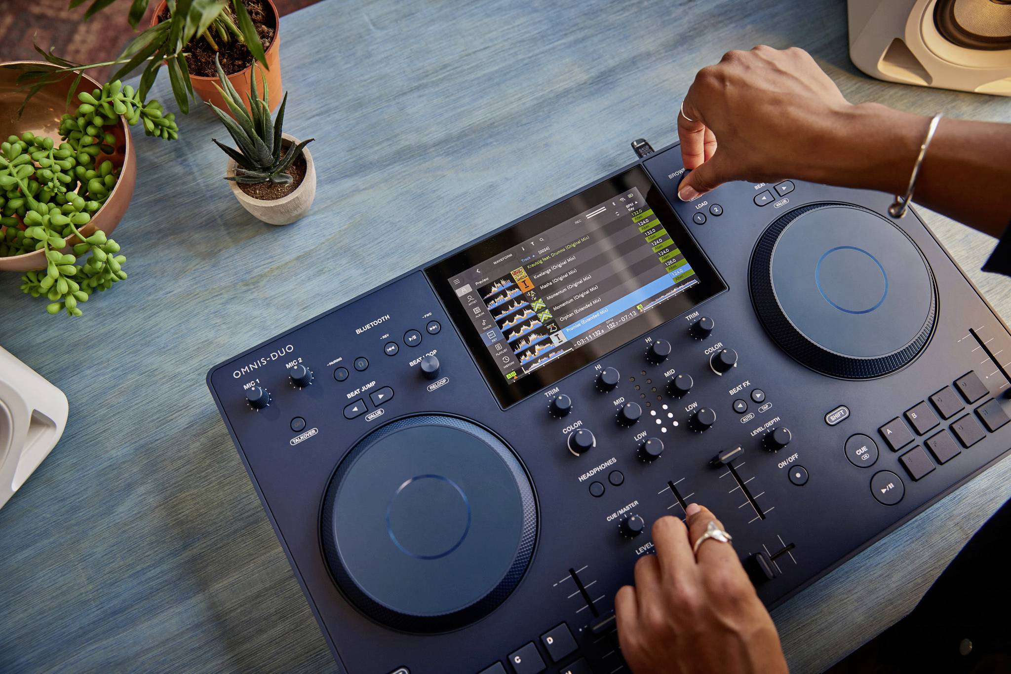 En person betjener et DJ-mixerbord med en skærm, der viser musiksoftware. Planter står i baggrunden på et træbord.