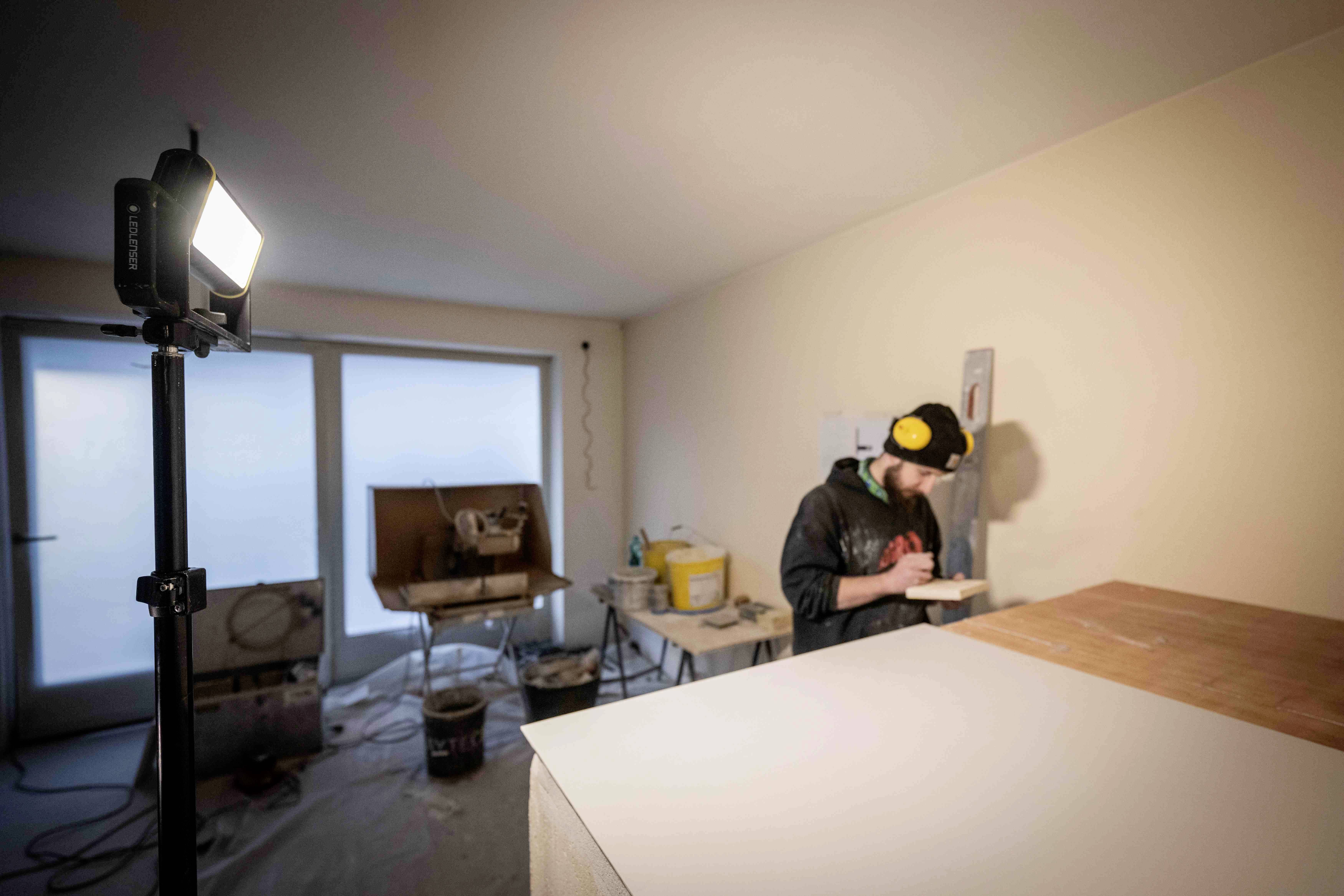 En håndværker arbejder på en pudset væg i et renoveret rum, omgivet af værktøjer og byggematerialer.