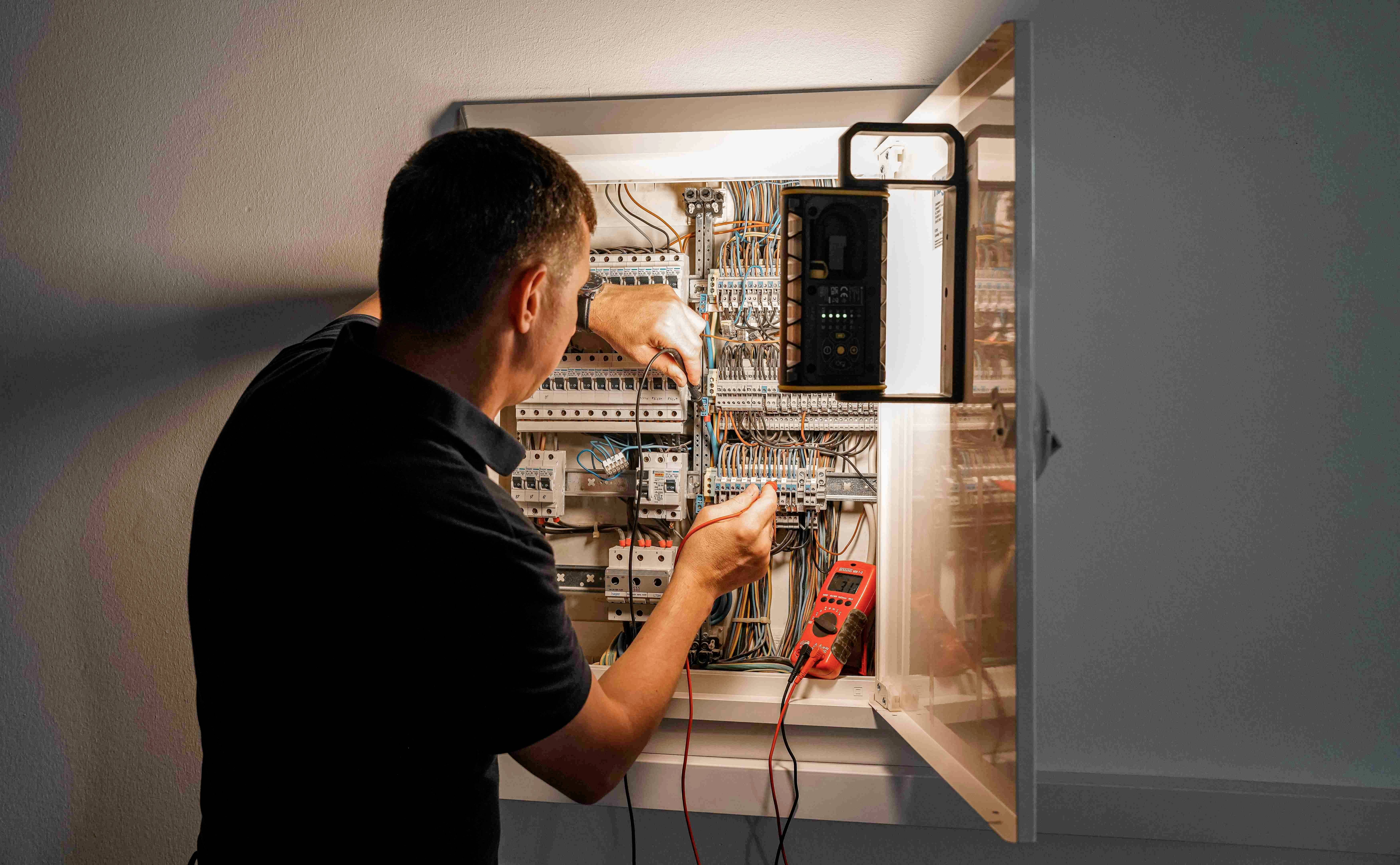 En mand arbejder ved en åben sikringsboks og kontrollerer kabelforbindelser med et multimeter til måling af elektriske værdier.