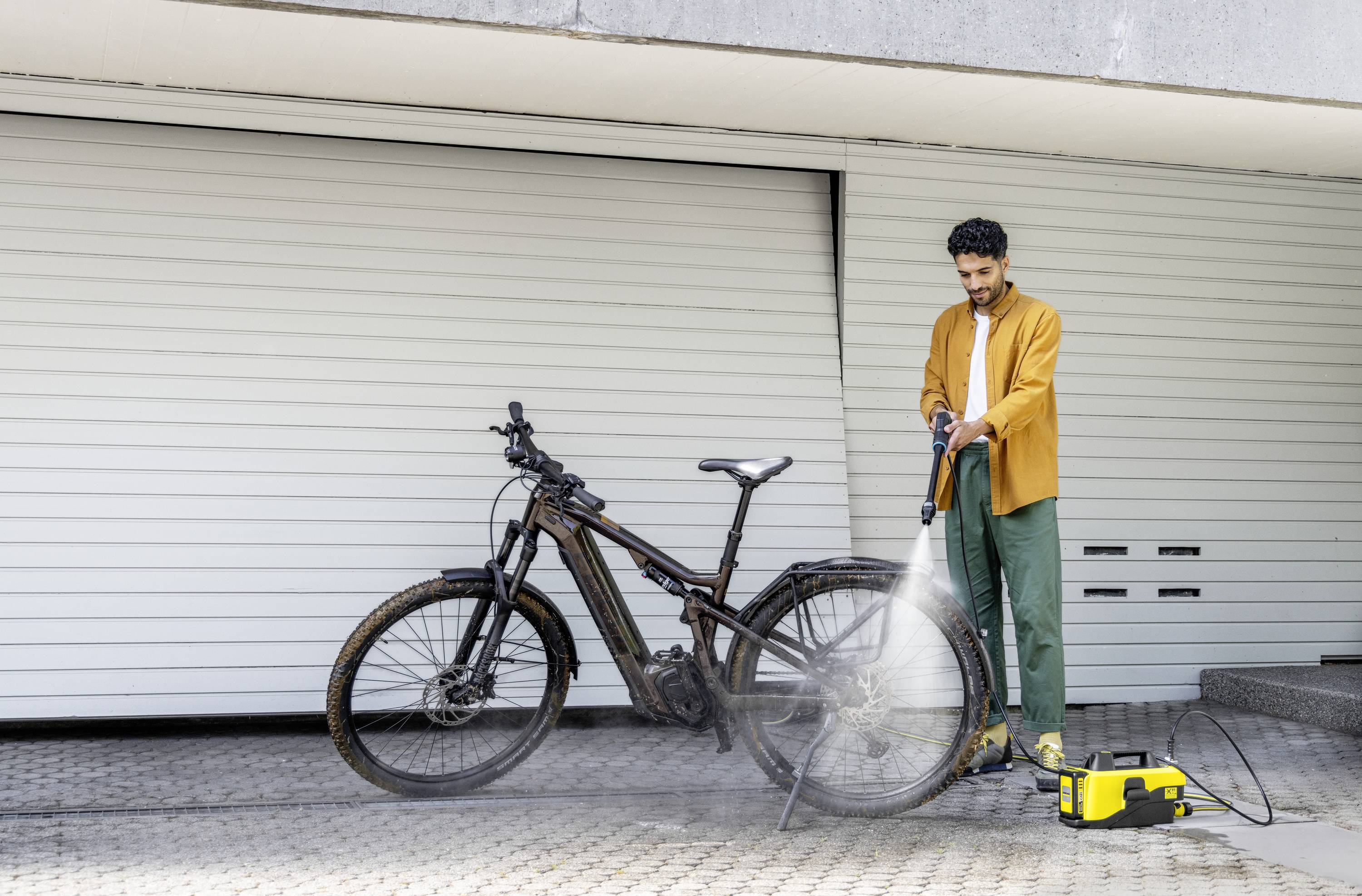 En mand rengør en beskidt cykel med en højtryksrenser foran en lukket garageport. Han bærer en orange jakke.