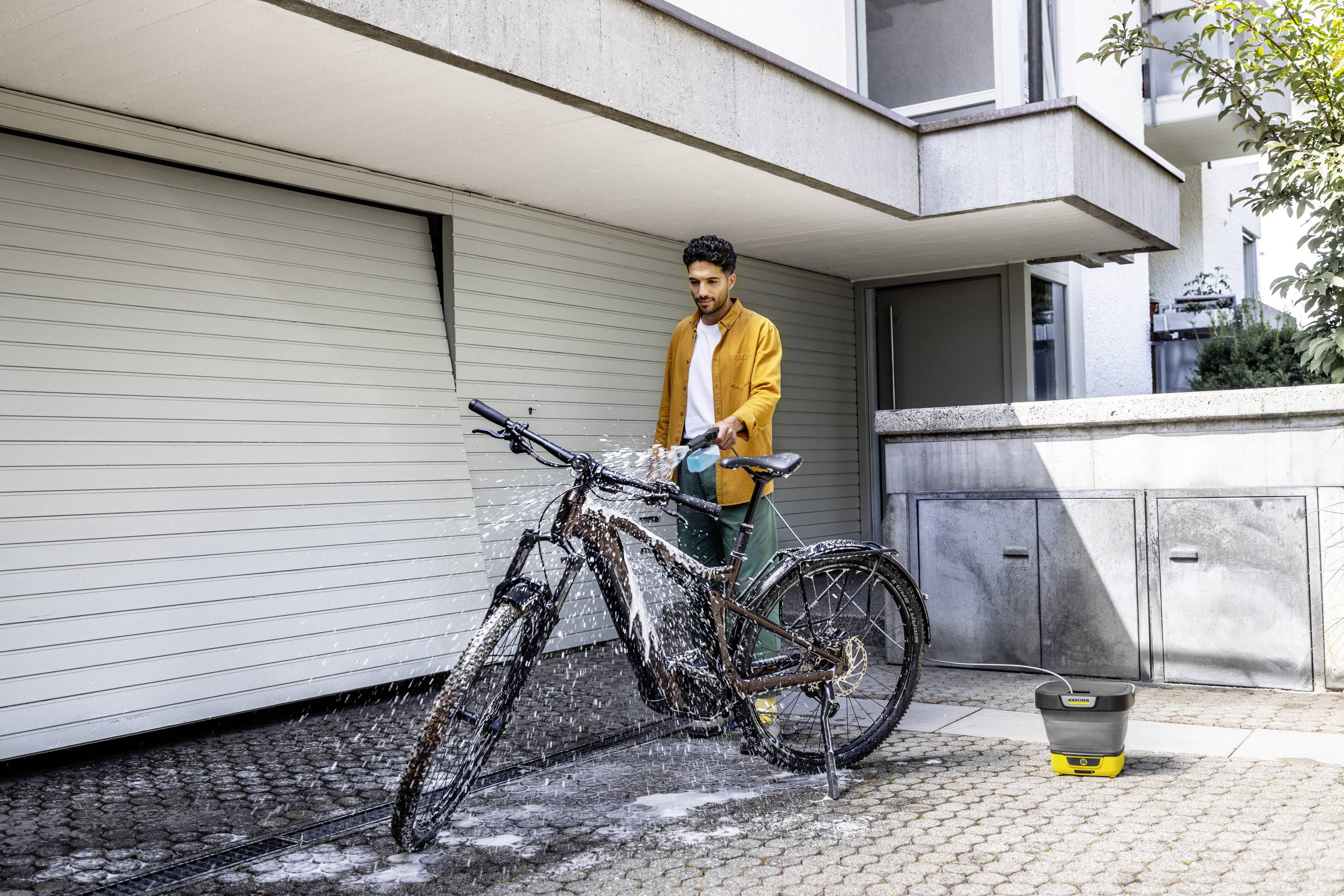 En mand i en gul jakke vasker sin cykel med vand på en brolagt plads foran en garage. En spand står ved siden af.