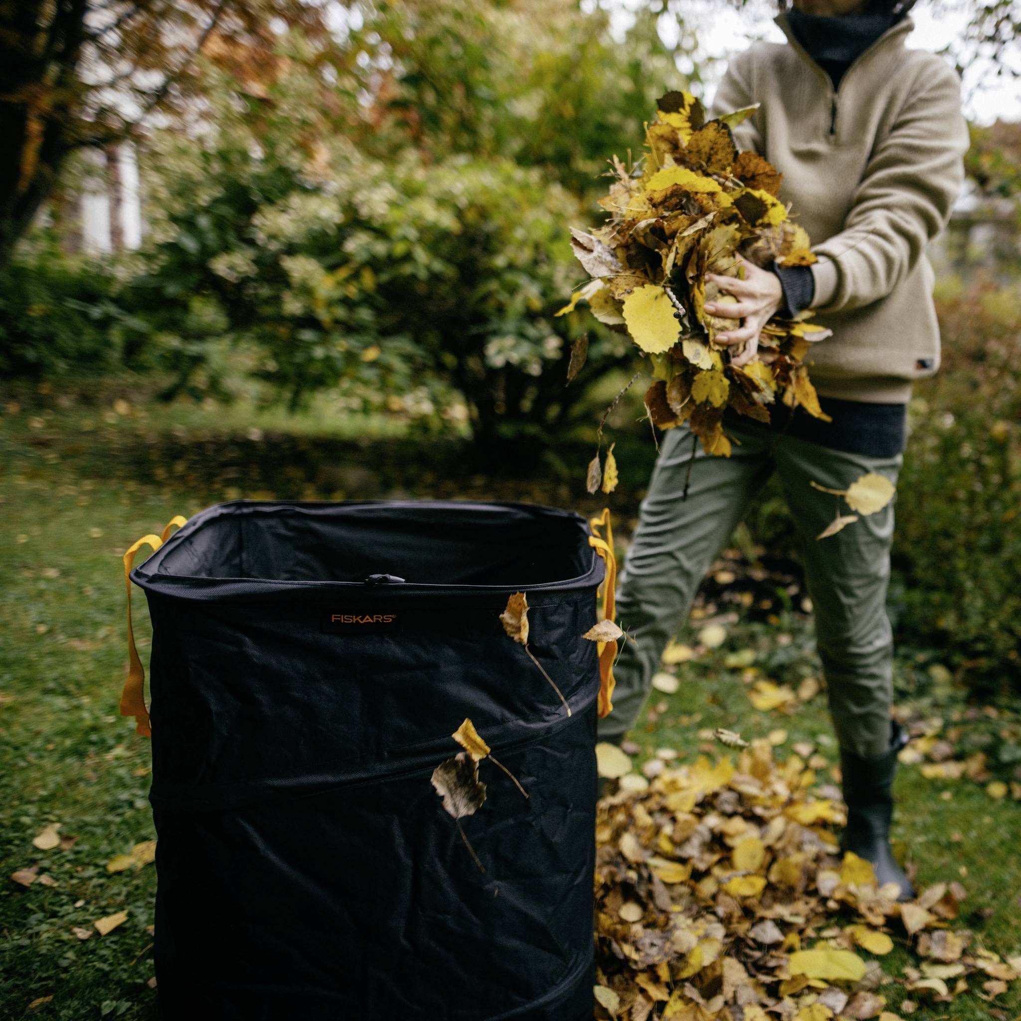 En person samler farverige efterårsblade og holder dem i hånden ved siden af en løvpose i en parkagtig have.