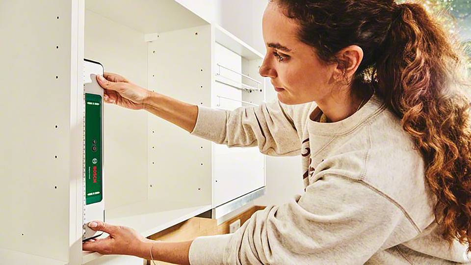 En kvinde måler en åben reol med en lasermåler. Hun bærer en grå sweater og er fokuseret på opgaven.