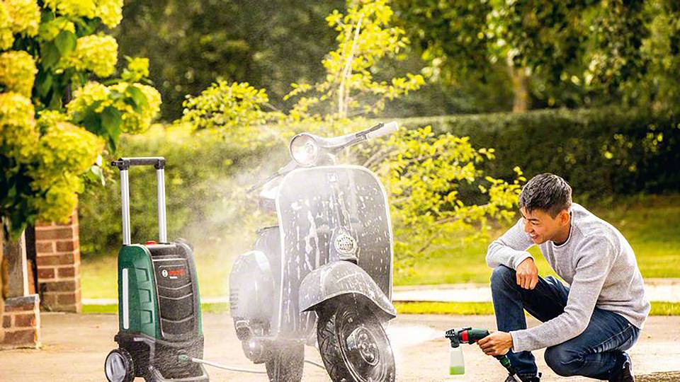 En mand rengør en scooter med en højtryksrenser udendørs. I baggrunden ses træer og buske.