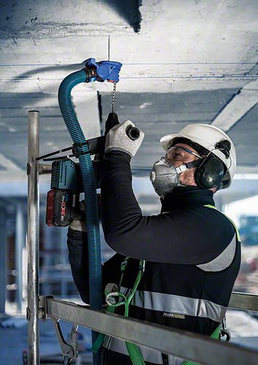 En arbejder i beskyttelsestøj og åndedrætsværn borer et hul i loftet. Han står på et stillads og bruger en elektrisk boremaskine.