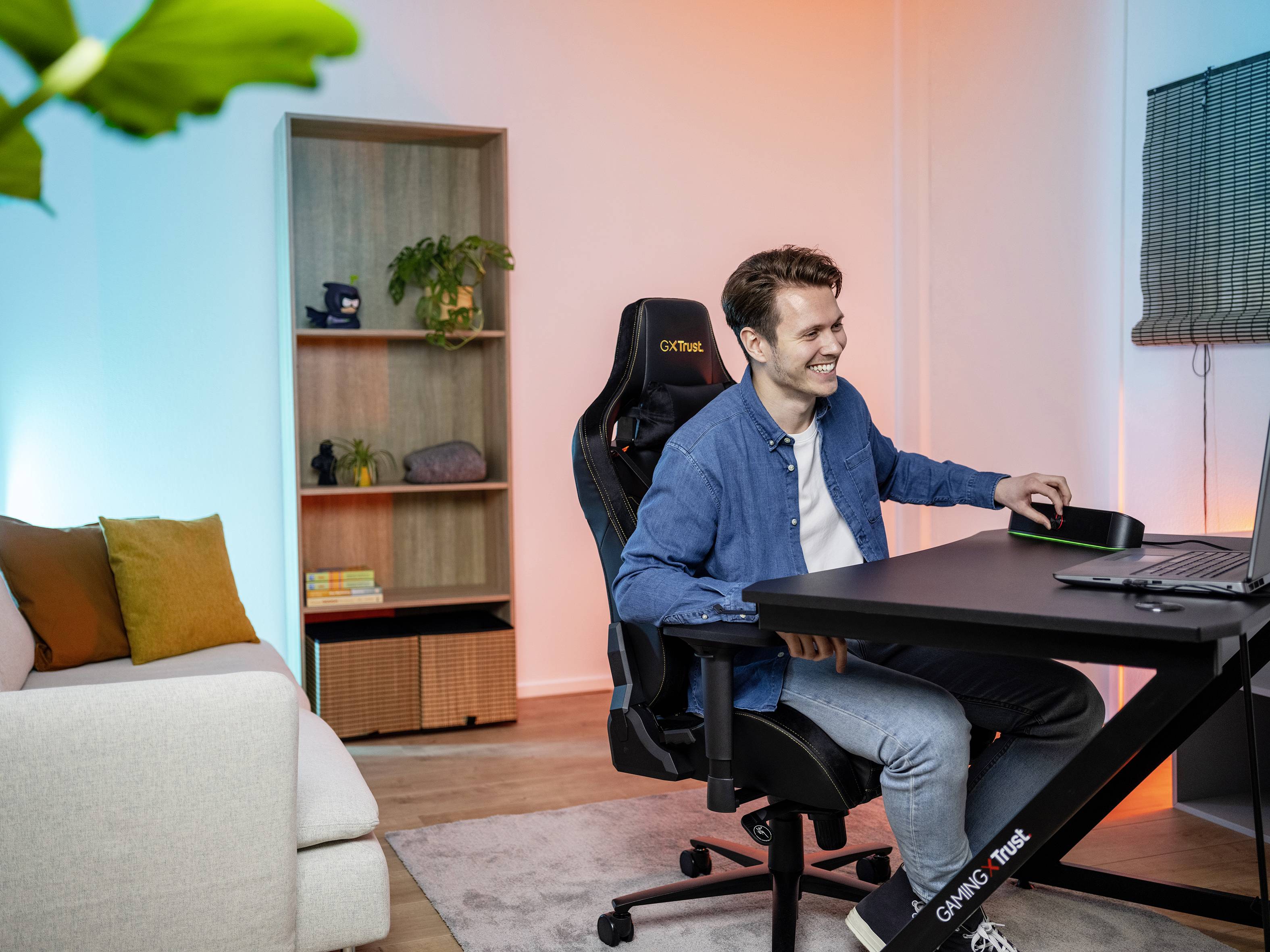 En mand sidder smilende ved et skrivebord med en computer, omgivet af moderne møbler. Atmosfæren virker hyggelig og fokuseret.