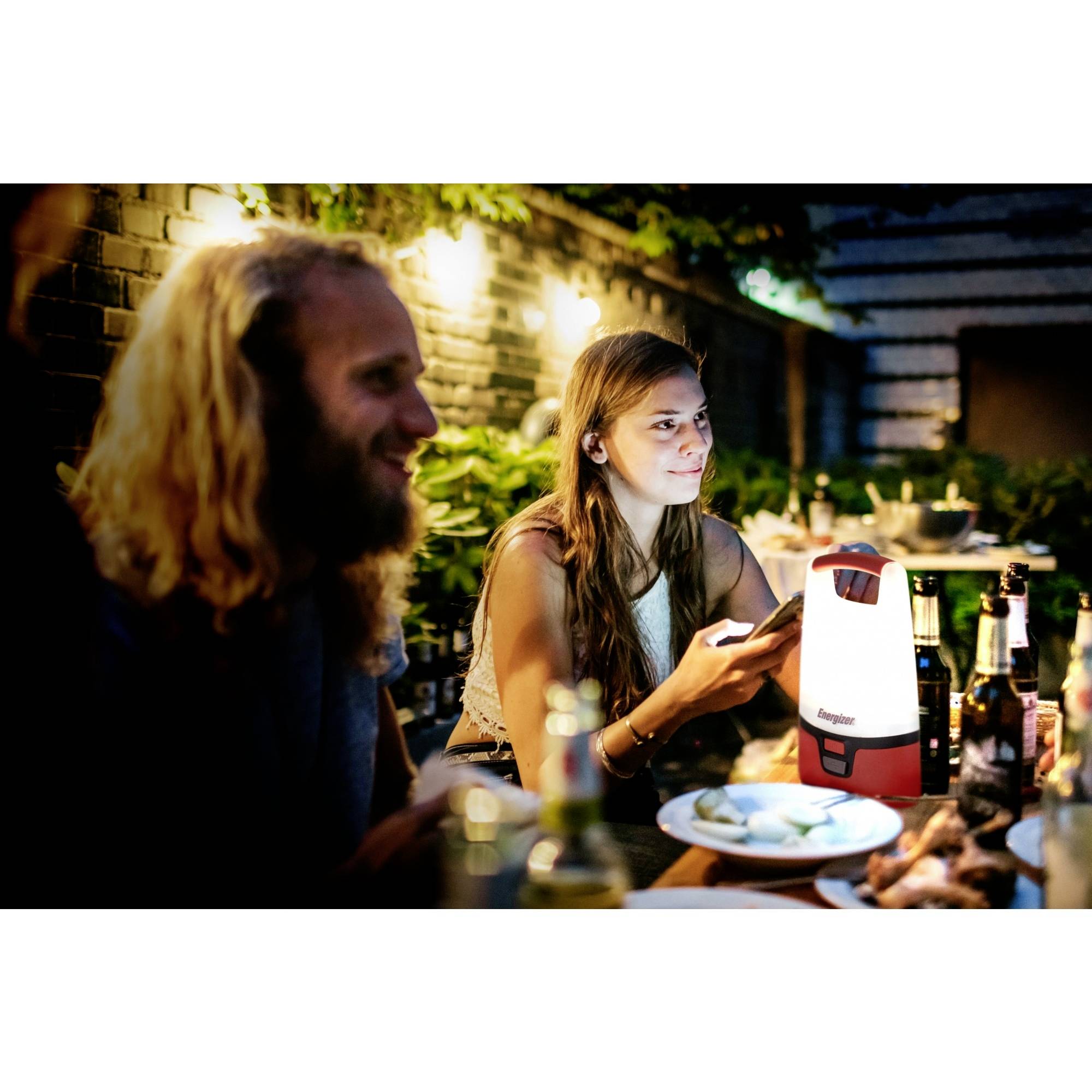 En gruppe mennesker sidder ved et udendørs aftensmåltid. En lysende lampe står på bordet. Der er en afslappet stemning.