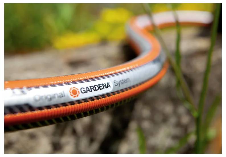 Nærbillede af en orange og grå GARDENA haveslange, der ligger på tværs af en stenede udendørs overflade, med sløret grønne områder i baggrunden.