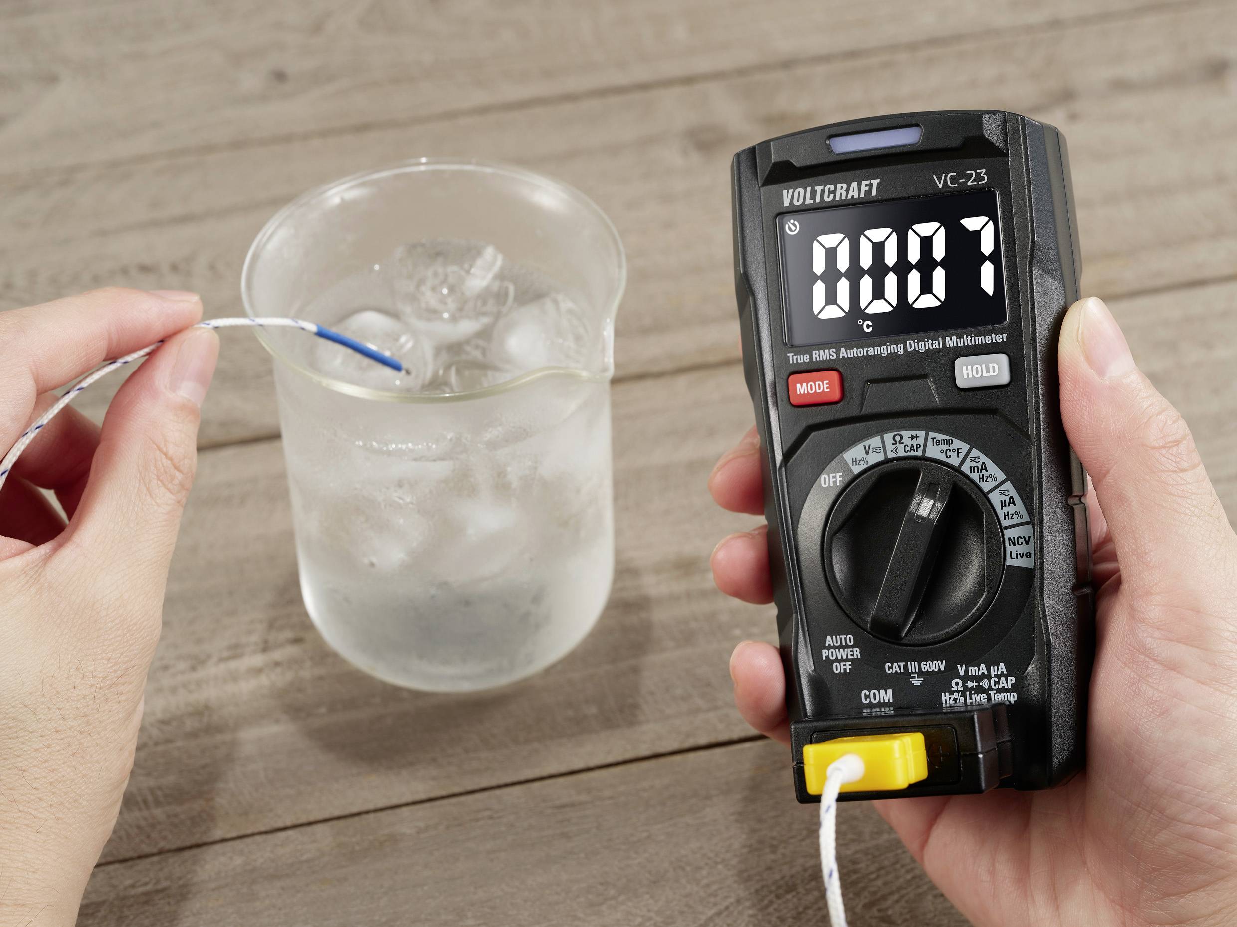 En person måler vandtemperaturen i et bægerglas ved hjælp af et digitalt multimeter, der viser 0,07 °C.