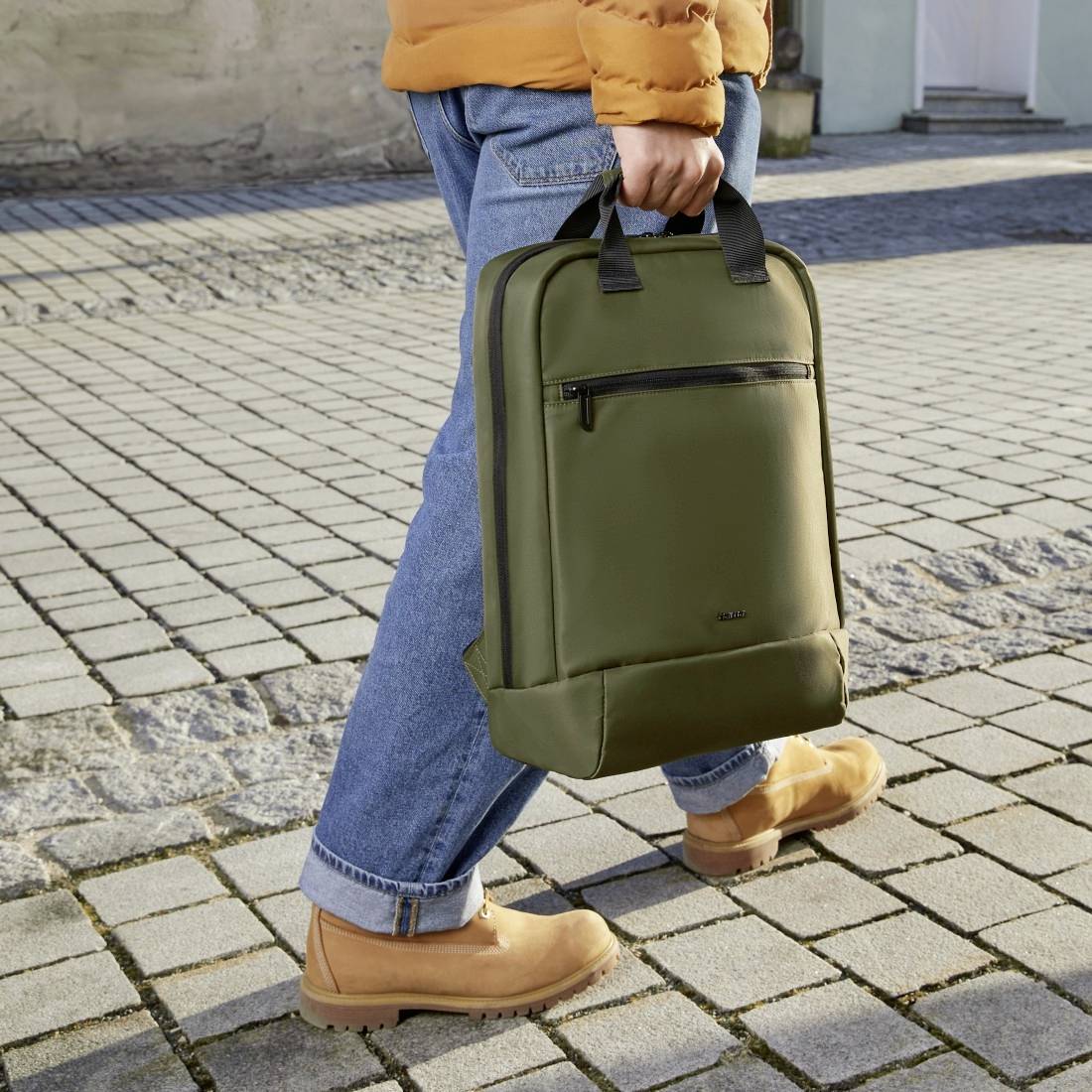 En person i orange jakke og blå jeans holder en grøn rygsæk, stående på en brolagt vej.