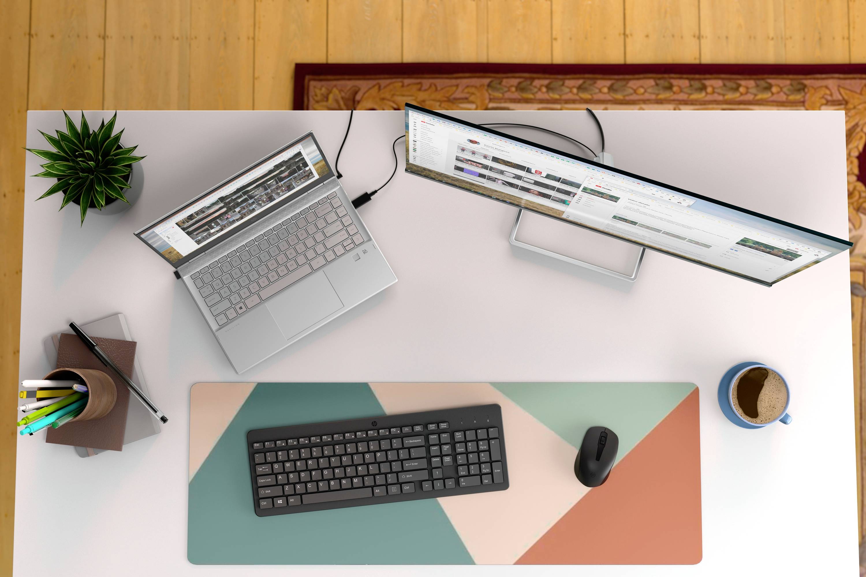 Et skrivebord med bærbar computer, skærm, tastatur, mus, plante, penneholder og en kaffekop, set oppefra.