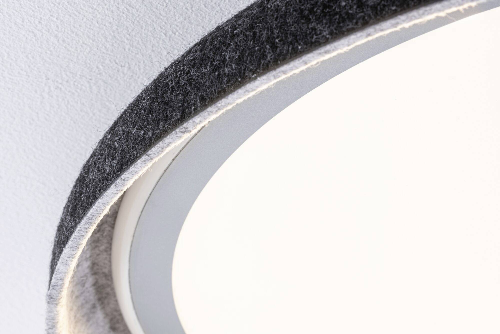 Del af en rund, moderne lampe på en hvid loftsoverflade, med en grå, filtlignende kant, der viser et raffineret design.