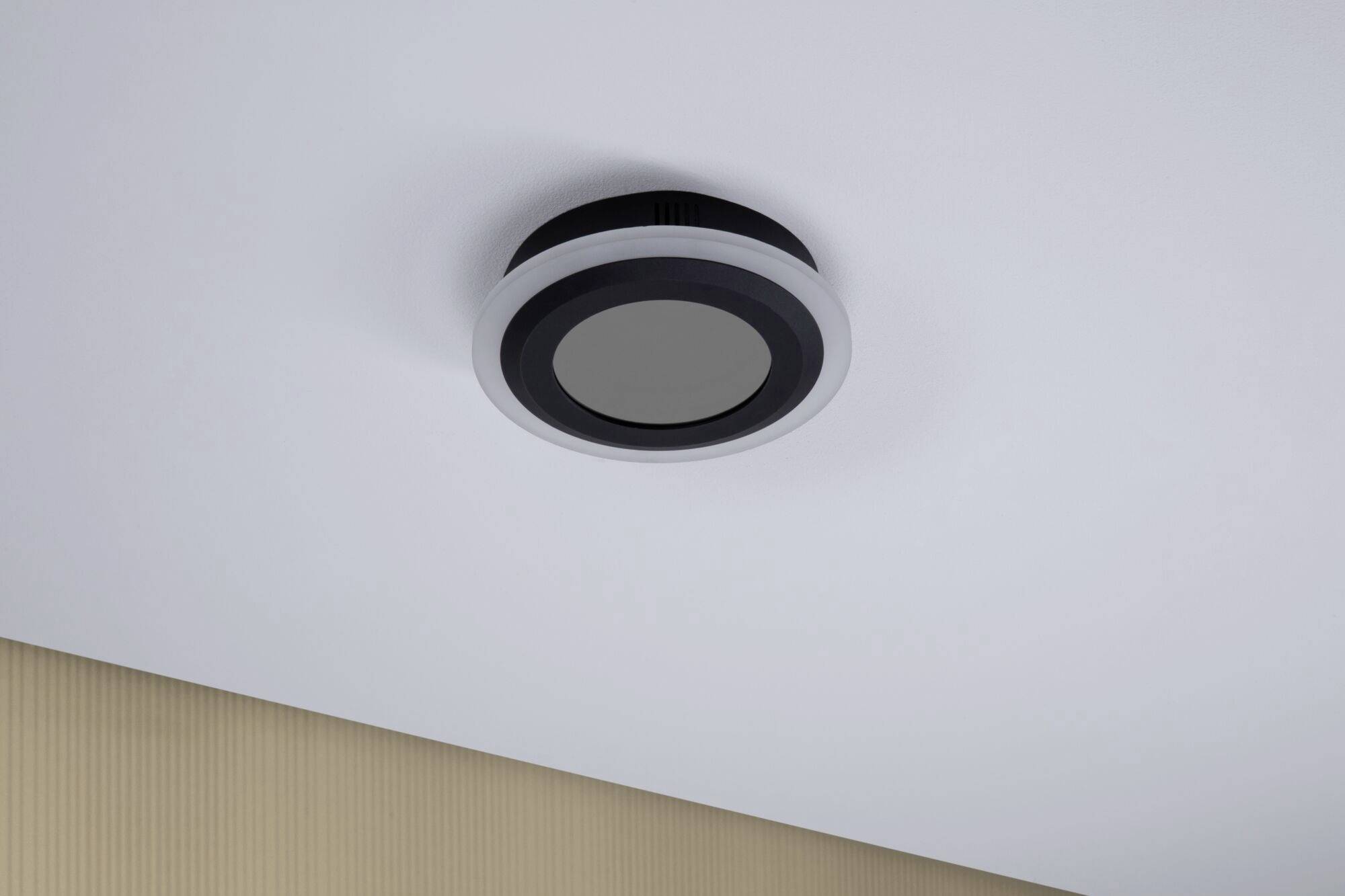 En rund røgdetektor er monteret på en hvid loft. Væggen nedenunder er beige. Enheden er holdt i et enkelt sort-hvidt design.