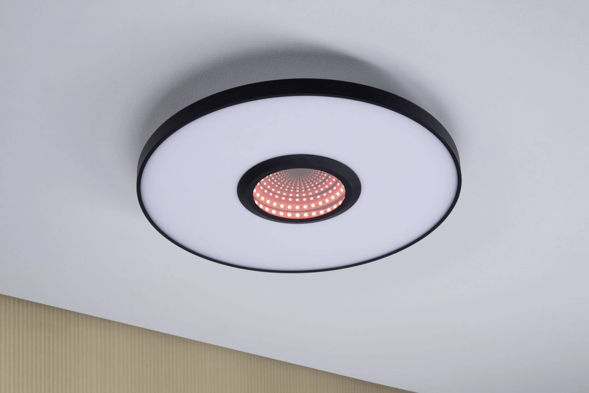 Rund loftslampe med LED, som lyser centralt rødt. Monteret på en hvid loft over et beige stribet vægparti.