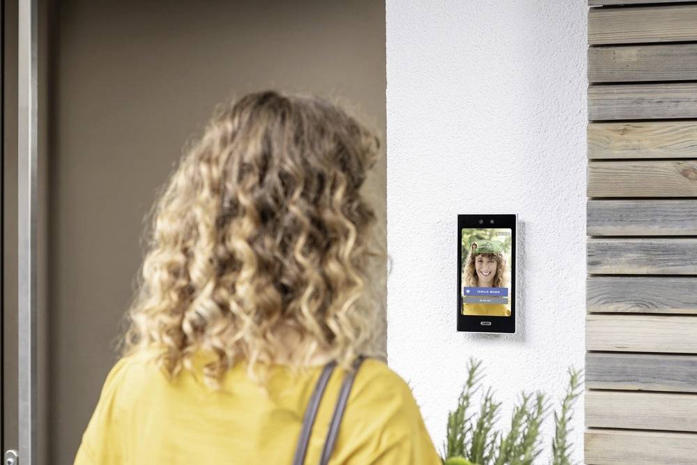 En kvinde med krøllet hår står foran en dørtelefonanlæg, som viser et billede af en person på skærmen.