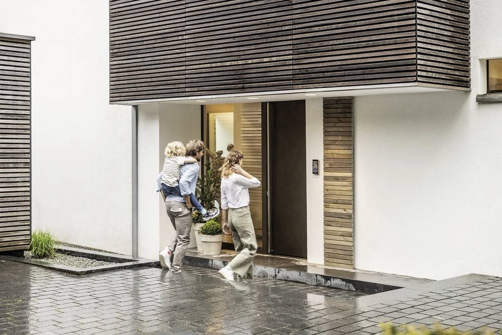 En familie træder ind i et moderne hus. En mand bærer et barn, kvinden åbner døren. Det regner let, og indgangspartiet er vådt.