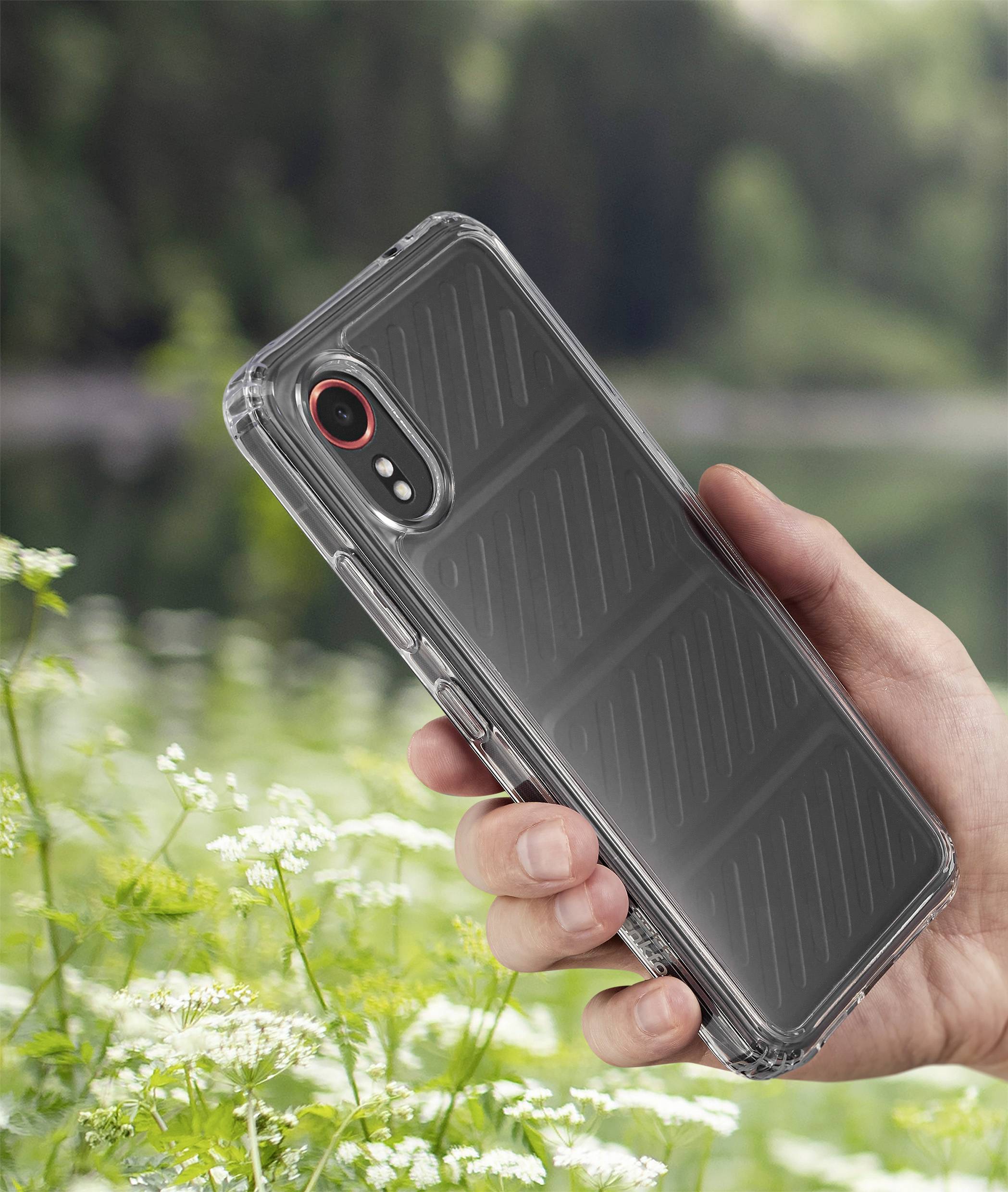 En hånd holder en smartphone med et gennemsigtigt cover foran et sløret, grønt skovlandskab med en sø i baggrunden.