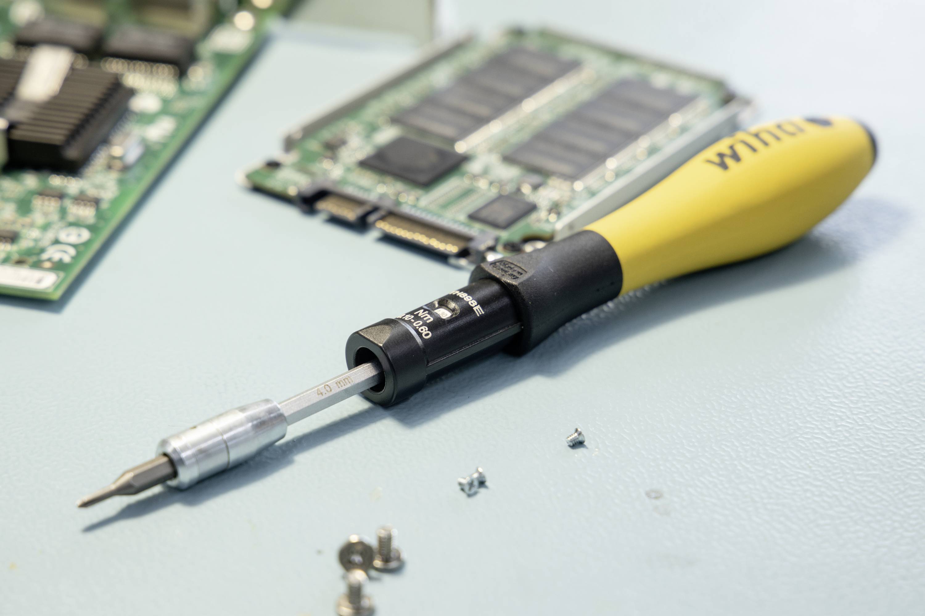 En gul skruetrækker ligger på et bord ved siden af computerkort og nogle små skruer. Den bruges til elektronikreparationer.