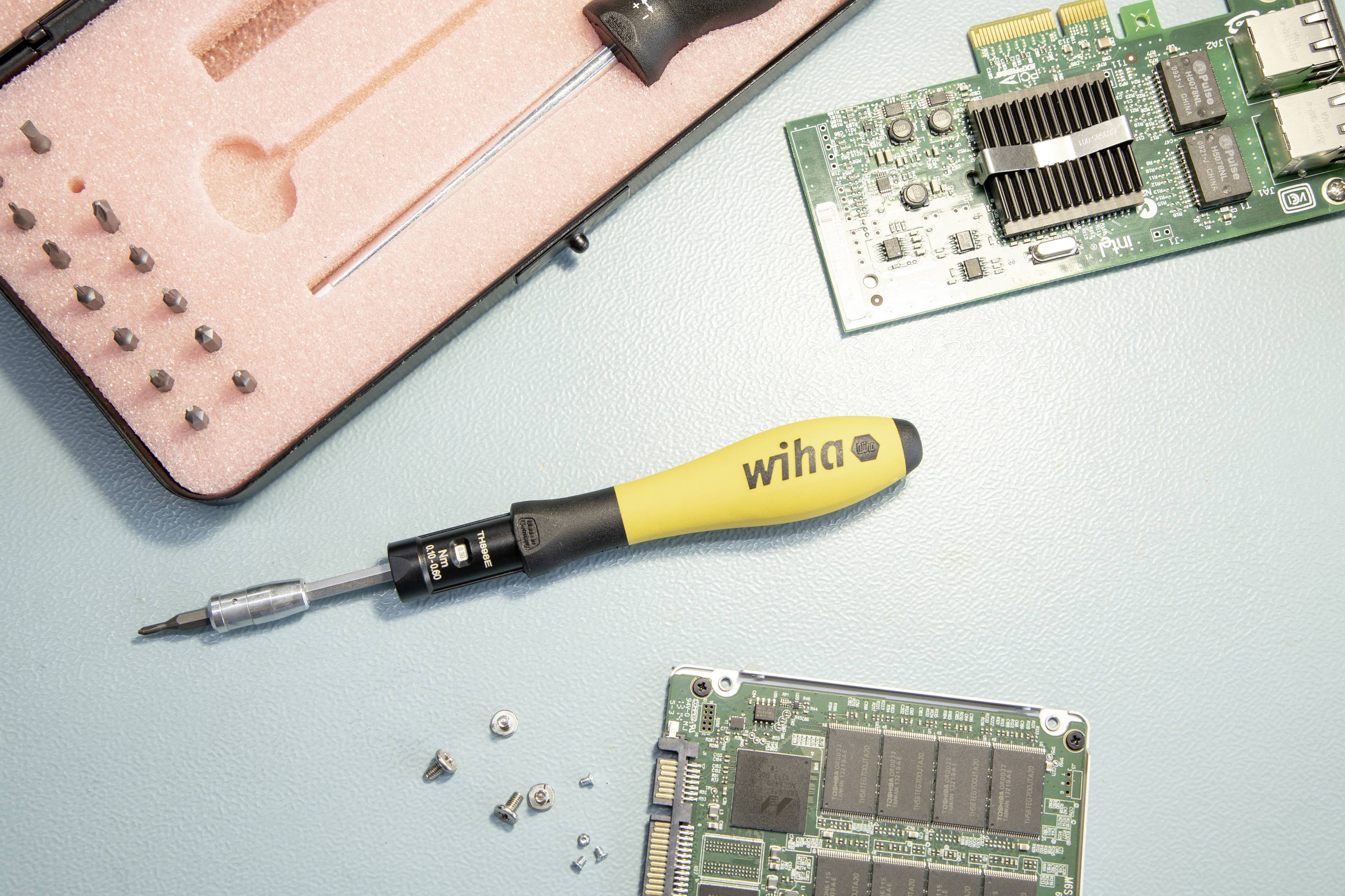 En gul skruetrækker ligger mellem computermotherboards og skruer på et lyseblåt bord. Et åbent etui med bits er synligt.