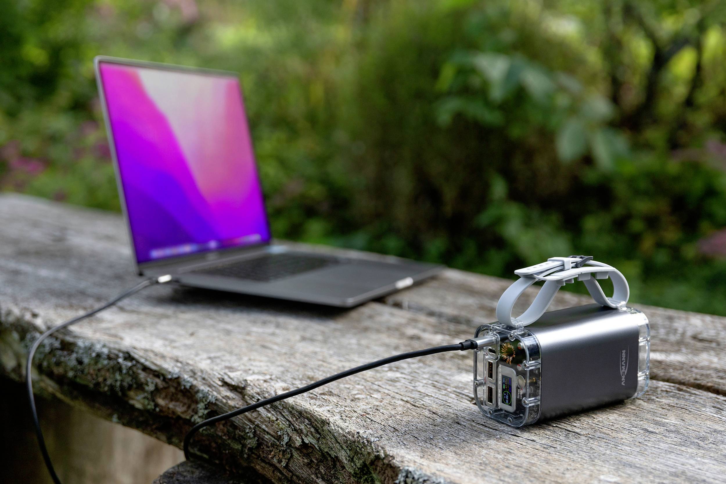 En bærbar computer står på et træbord udendørs, forbundet med en powerbank, der ligger ved siden af den, omgivet af planter i baggrunden.