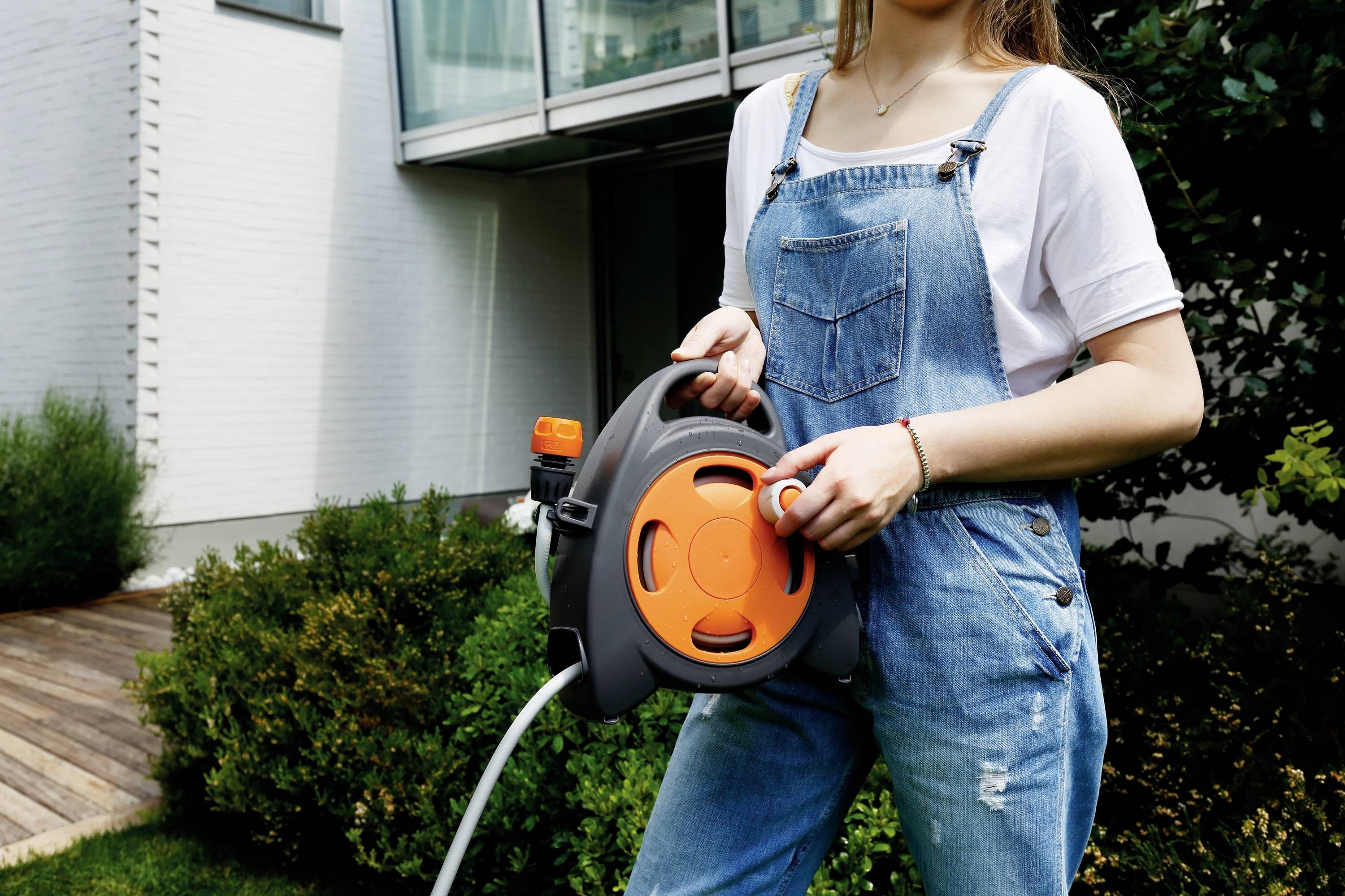 En person i jeans-overall holder en orange haveslange-oprulningsanordning foran en moderne bygning med have.