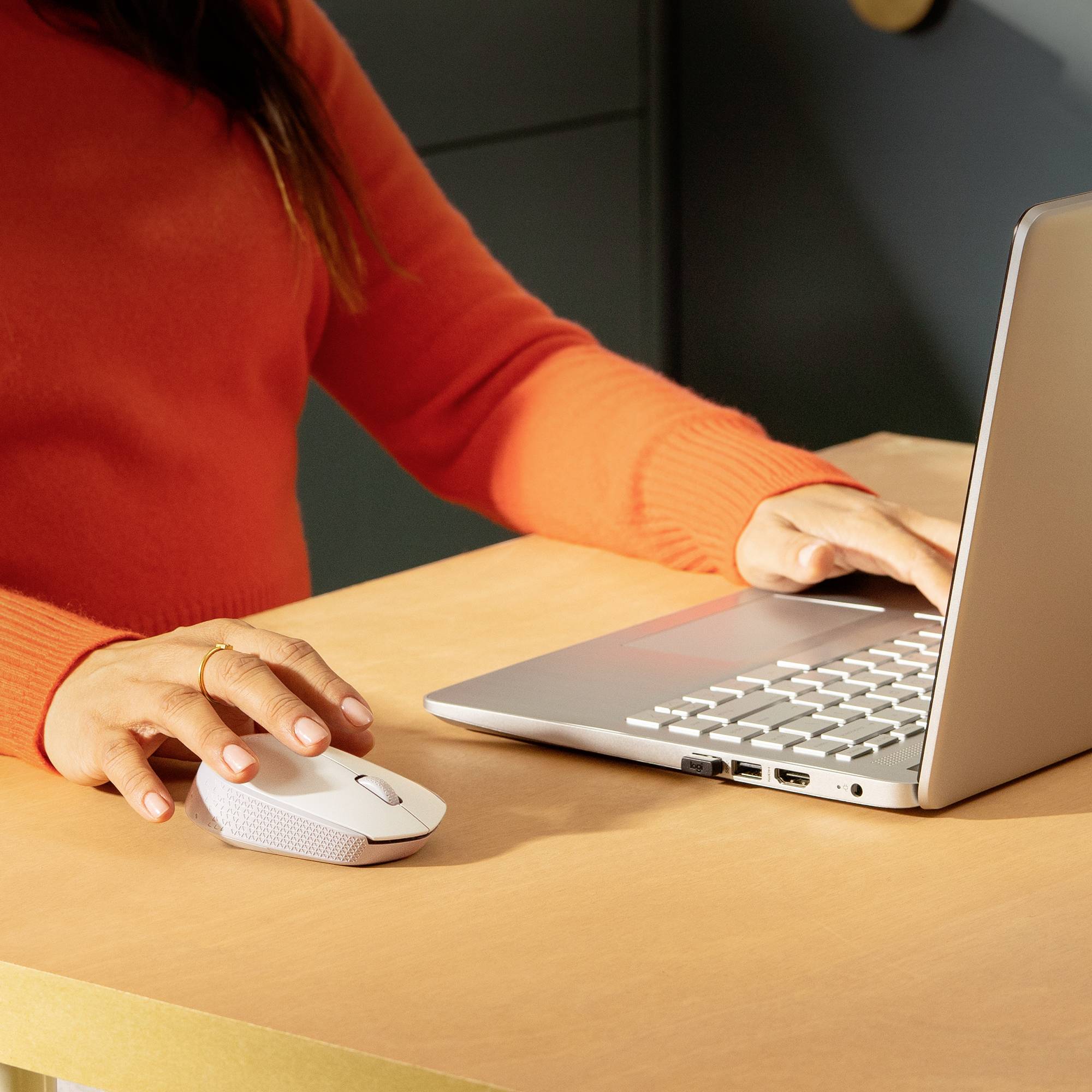 En person i en orange trøje bruger en bærbar computer og en trådløs mus på et bord. Teknologi og arbejde i fokus.