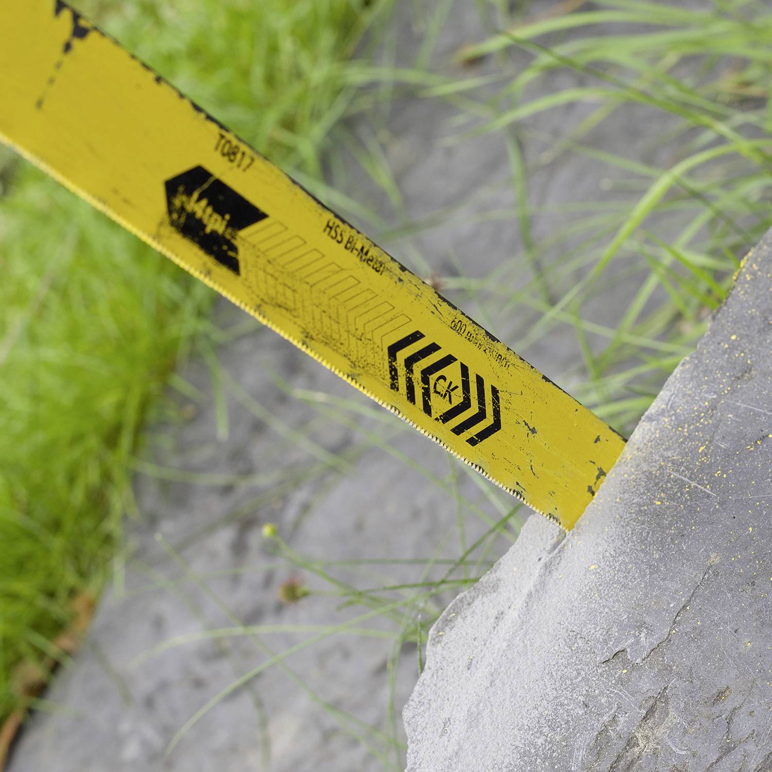 Et gult målebånd ligger på en sten, omgivet af græs. Målebåndet viser centimetermærkninger og har sorte pile som design.