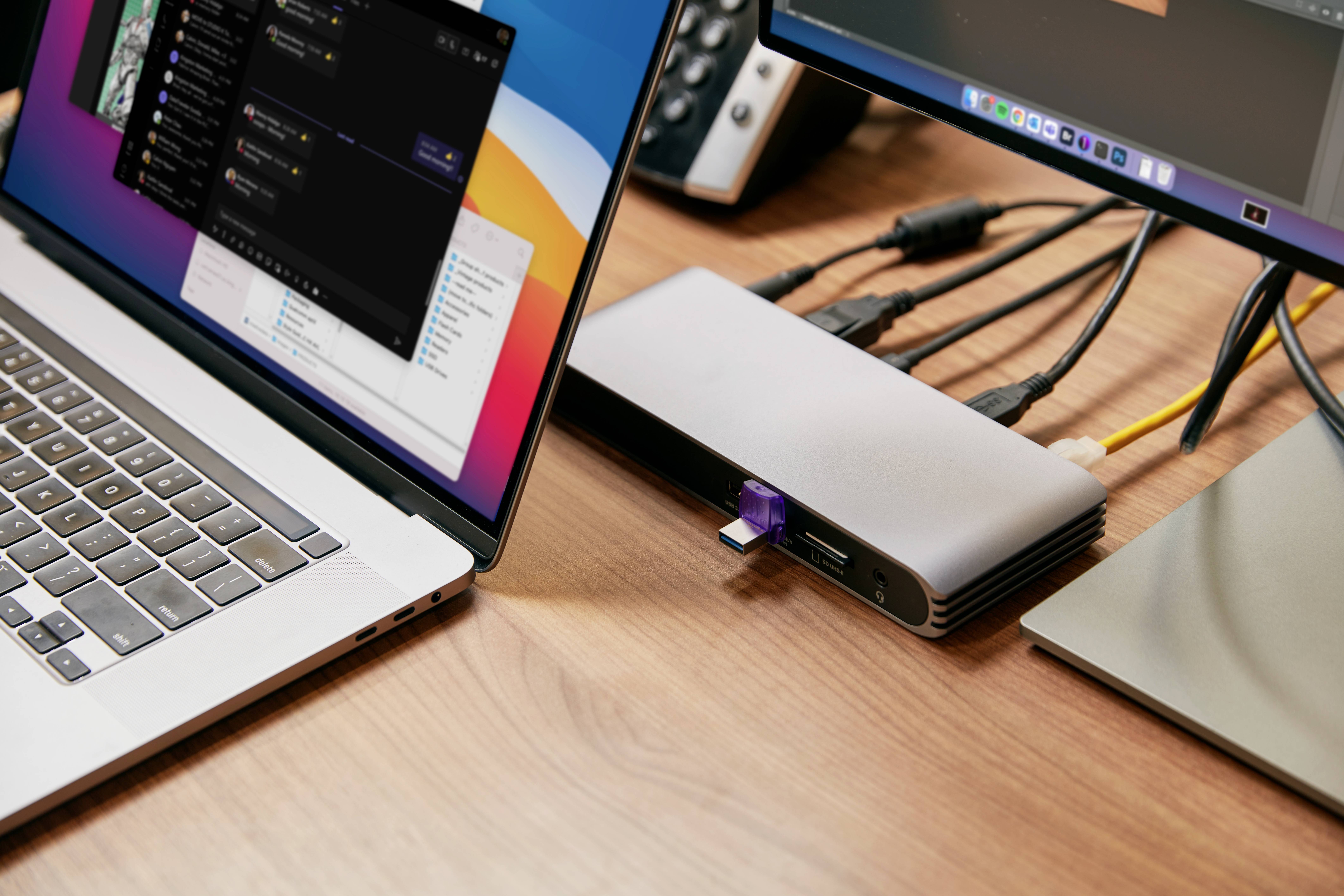 Bærbar computer på et skrivebord, forbundet med flere enheder via en USB-hub. Skærmen viser åben software. Kontorarbejdsmiljø.