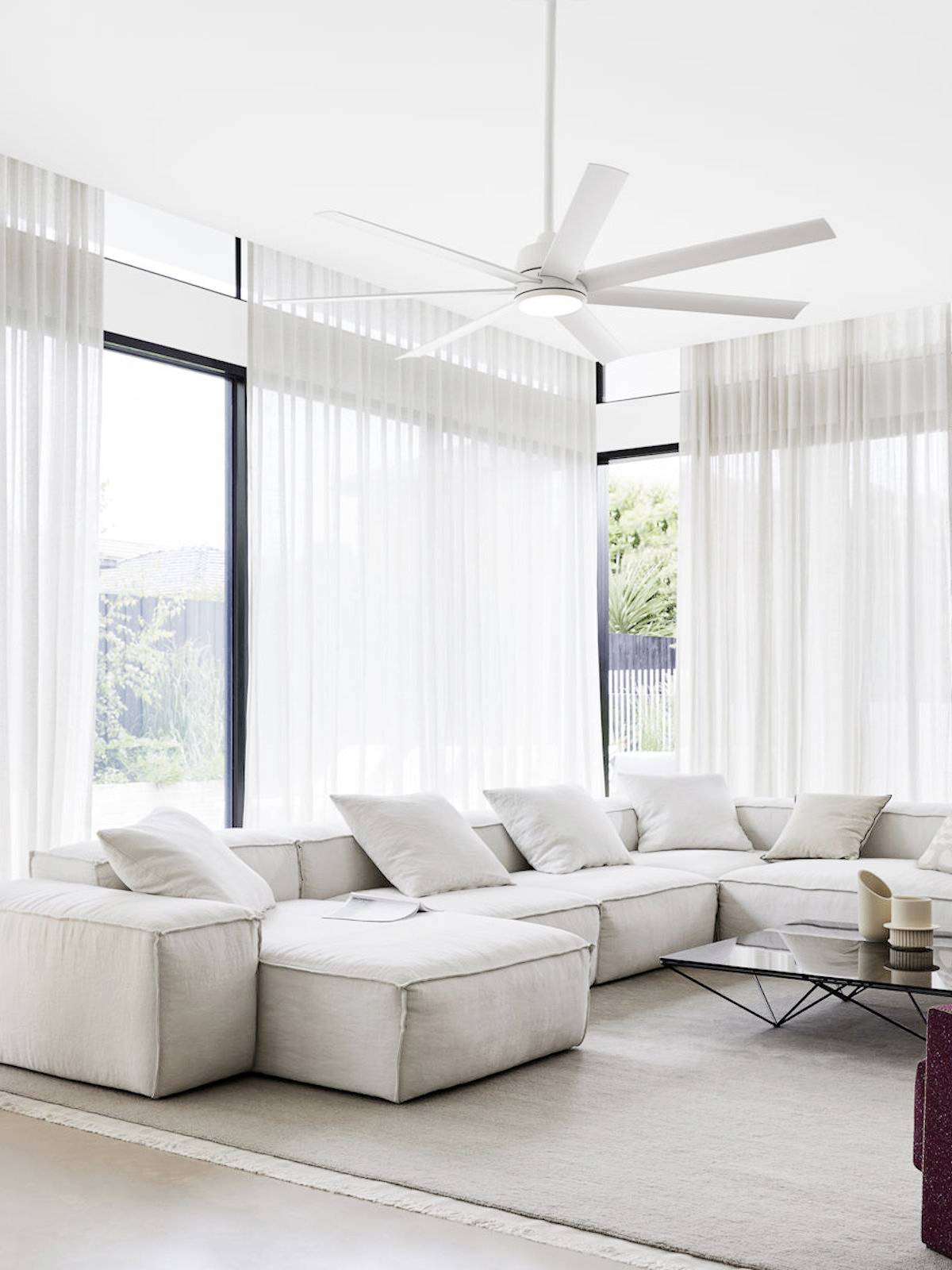 Et moderne wohnzimmer med store vinduer, en hvid hjørnesofa, et glasmesebord og en loftsventilator, lys og luftig atmosfære.