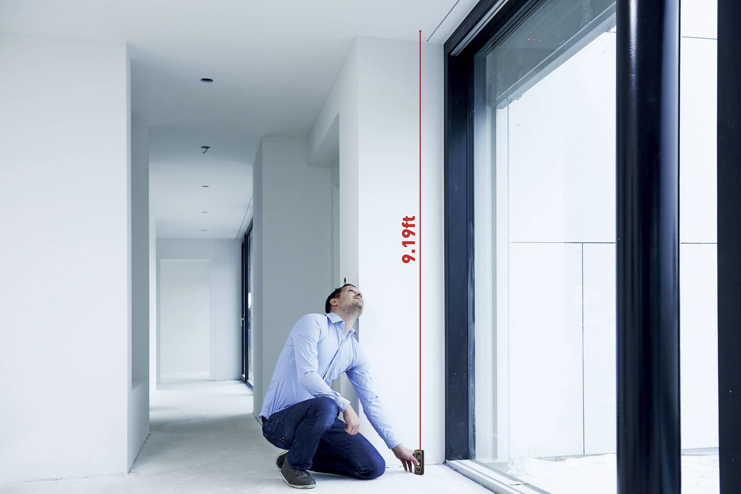 En mand knæler i et lyst rum og måler højden af et stort vindue med et målebånd. Højden er 9,19 fod.