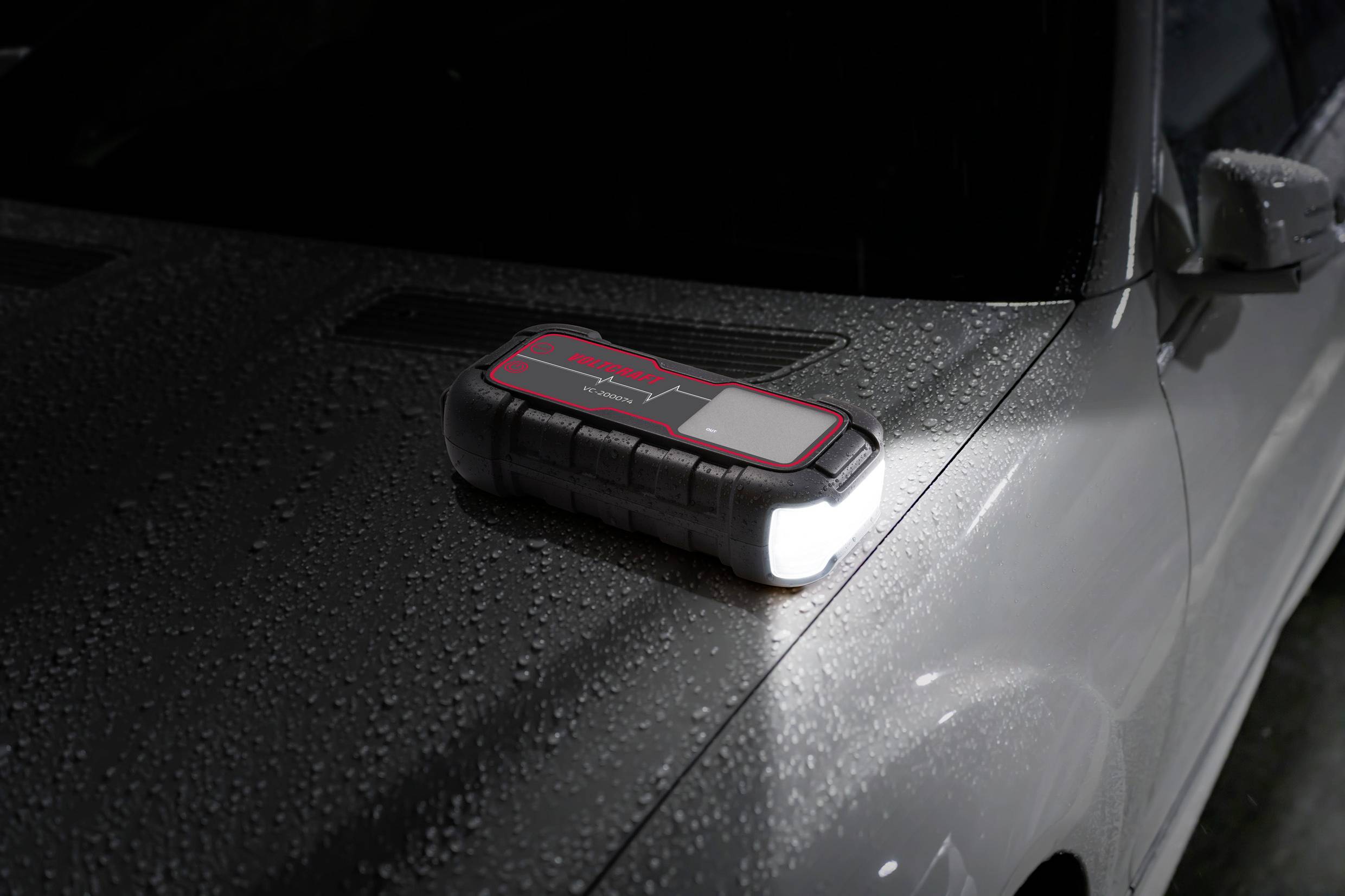 En sort bærbar powerbank med lommelygte ligger på motorhjelmen af en våd bil.