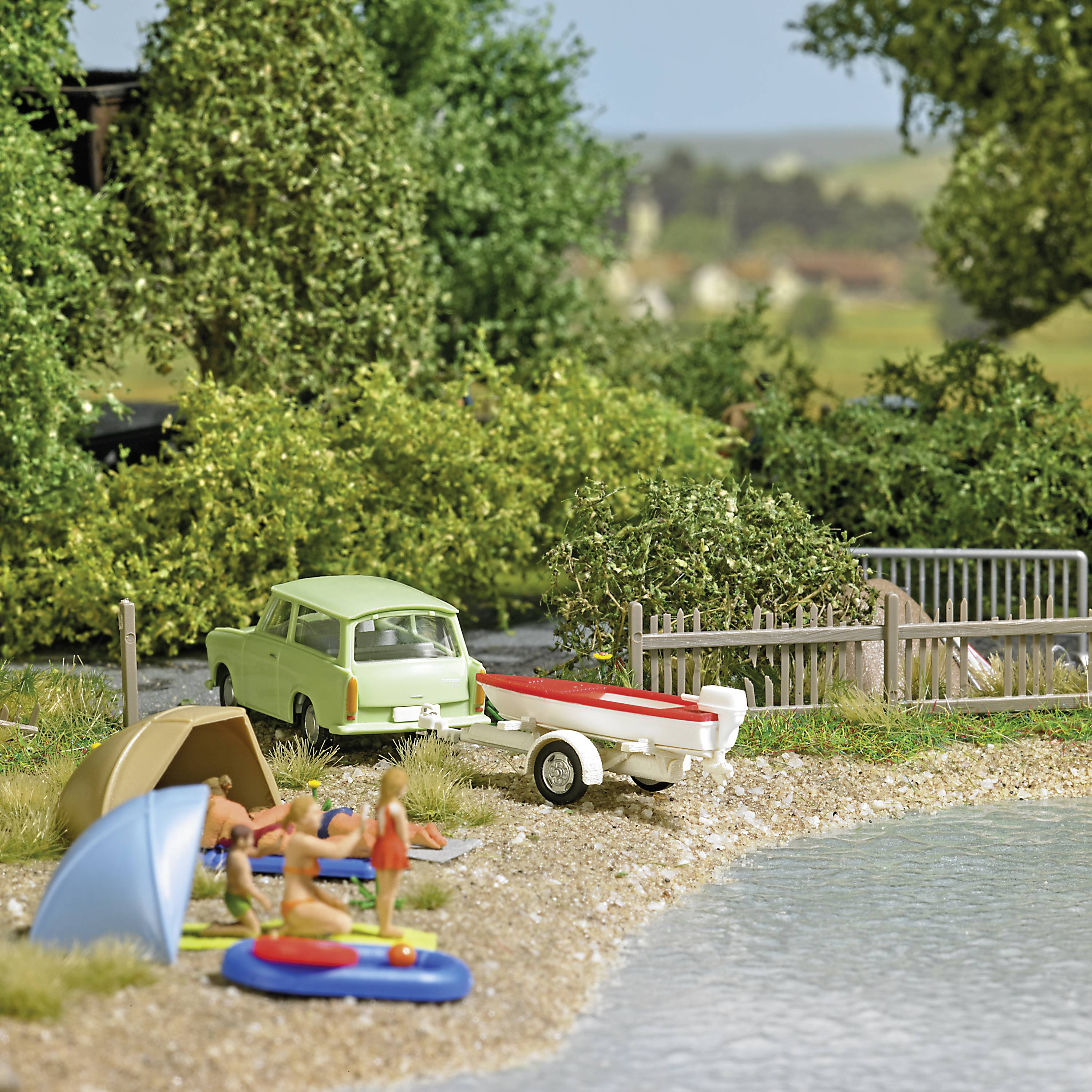 En miniaturemodel viser en campingscene ved søen med telte, en bil, en båd og mennesker, der slapper af. Baggrund: grønt landskab.